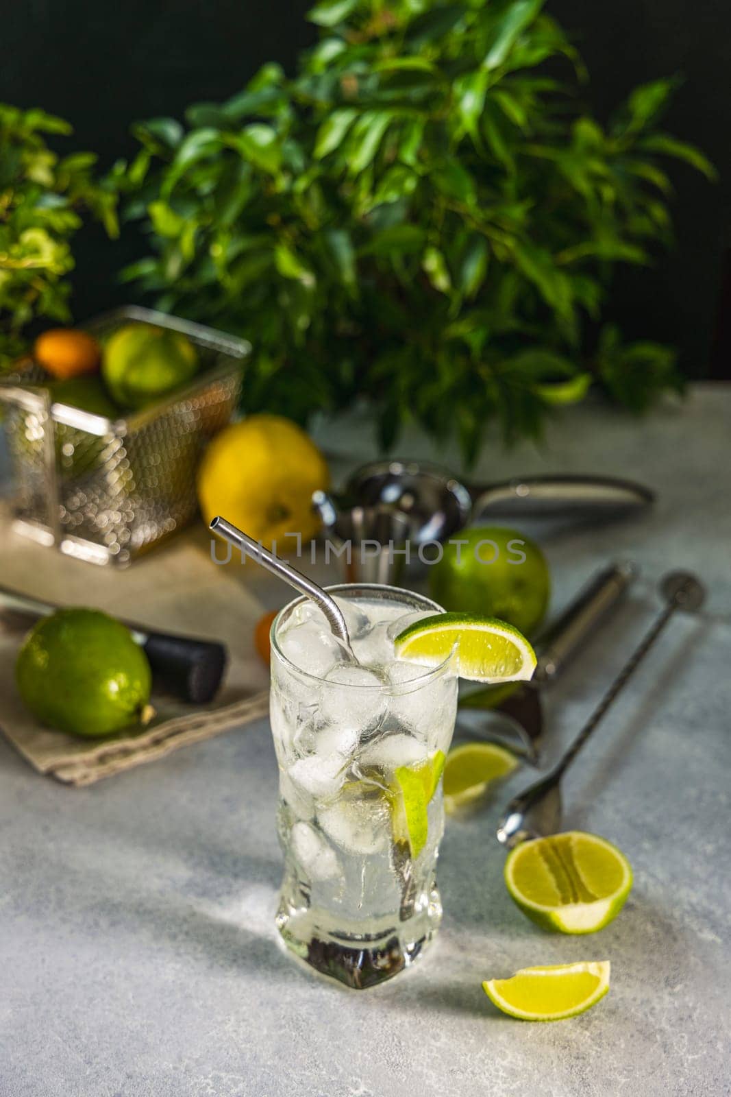 Gin and tonic cocktail being prepared with fresh lime, ice cubes, and metal straw, surrounded by bar tools and ingredients, ready for happy hour