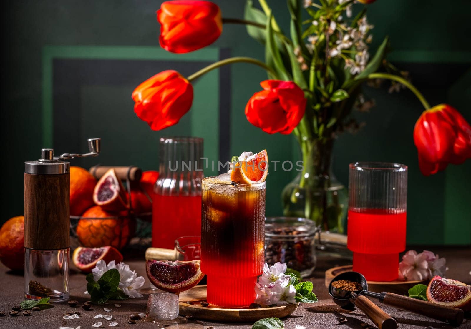 Glass of iced coffee with blood orange juice decorated with slice of orange and mint leaves, grinder, red tulips and ingredients on table