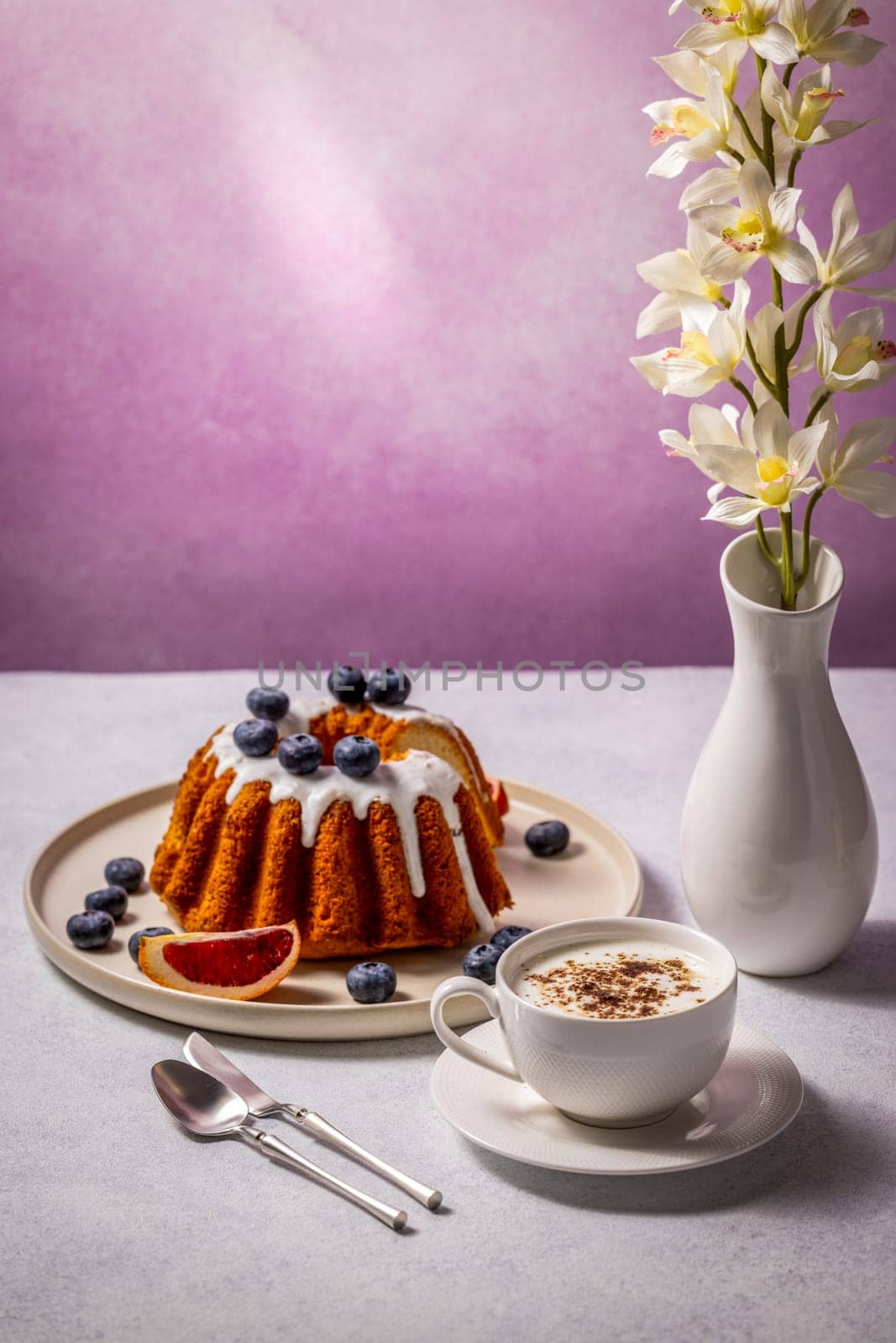 Round shaped cake with icing and blueberries served on a plate with a slice of blood orange together with a cup of cappuccino, perfect for a sweet breakfast