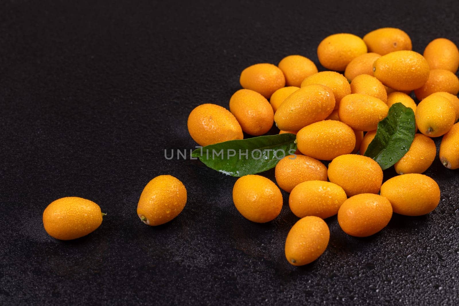 Vibrant kumquats, adorned with water droplets and accompanied by green leaves, create a striking contrast against a dark, wet backdrop