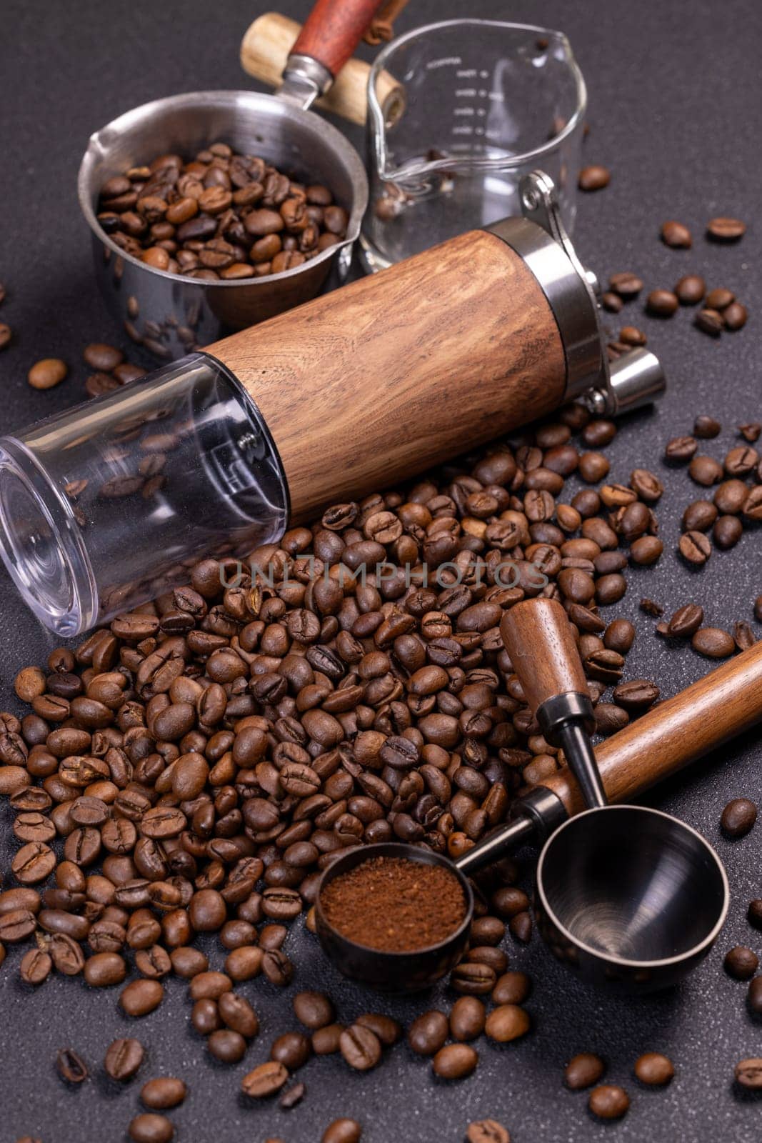 Roasted coffee beans scattered on a dark surface, accompanied by a manual grinder, metal scoop, and a measure of ground coffee, creating an aromatic tableau