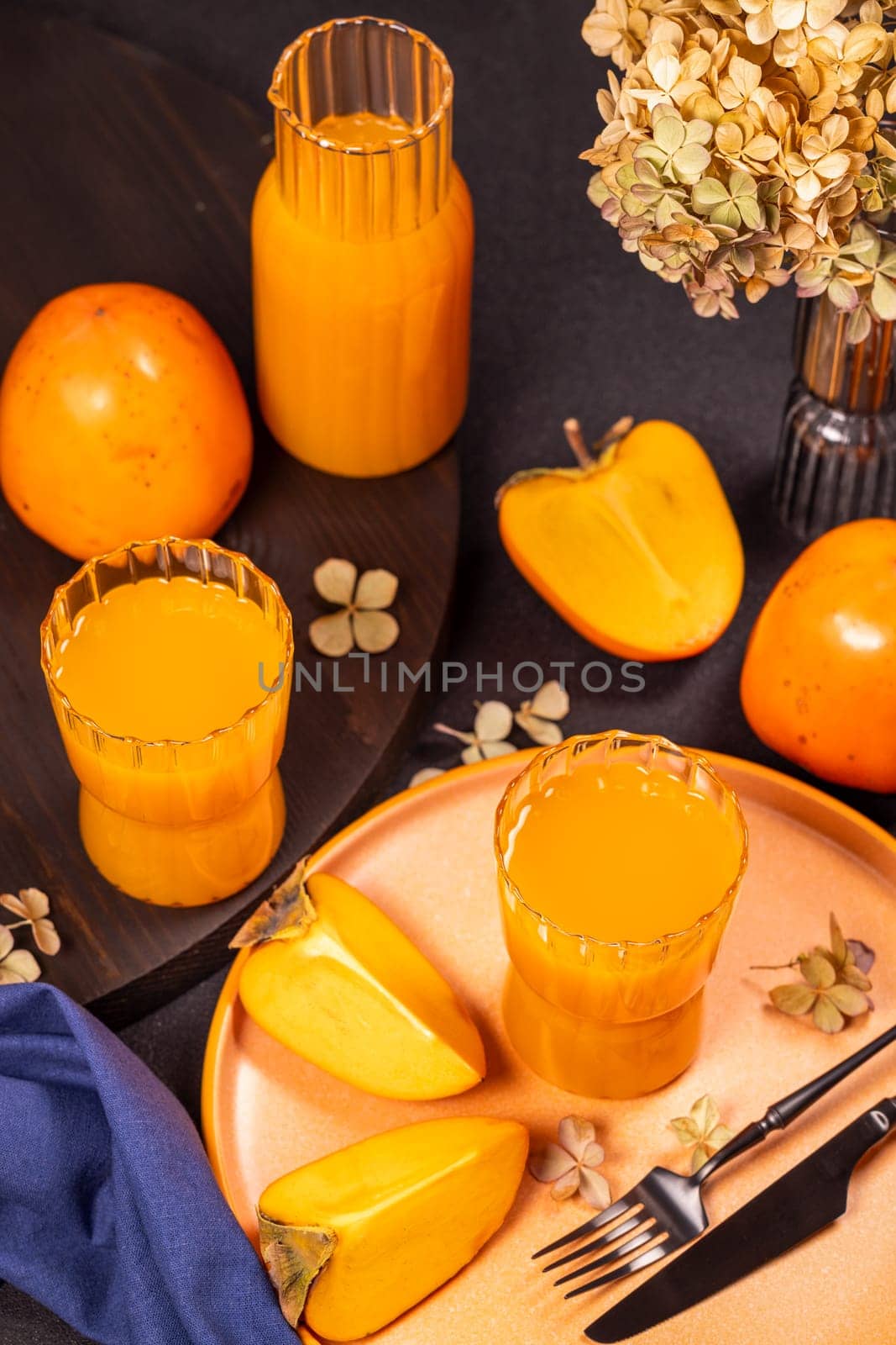 Two glasses and a carafe of persimmon juice are surrounded by fresh fruits and dried flowers, creating a vibrant and autumnal scene