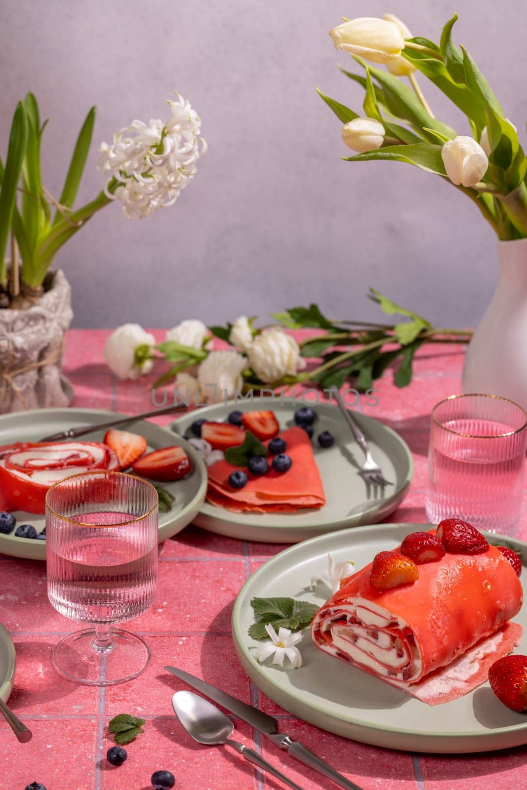 Delicious pink strawberry crepes and rolls served on green plates on a pink tiled table decorated with spring flowers and glasses of water