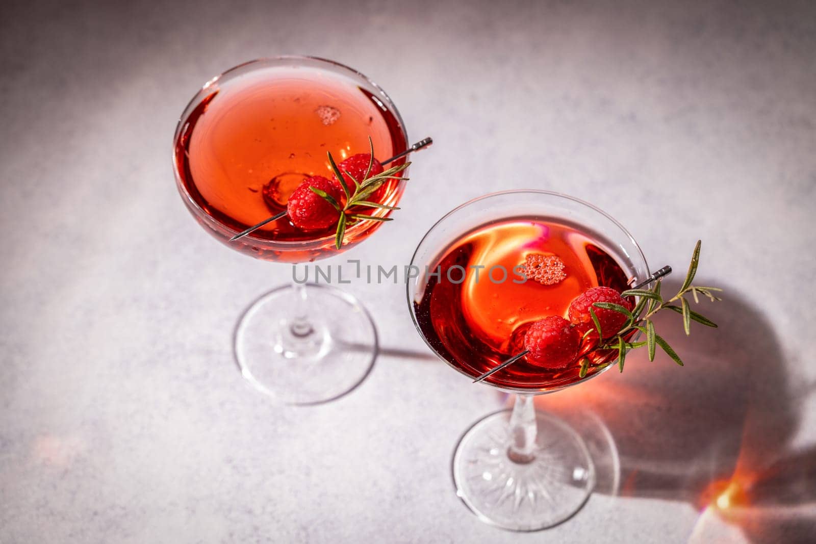 Two refreshing cocktails garnished with fresh raspberries and rosemary sprigs, served in elegant glasses on a grey table, creating a stylish and inviting scene