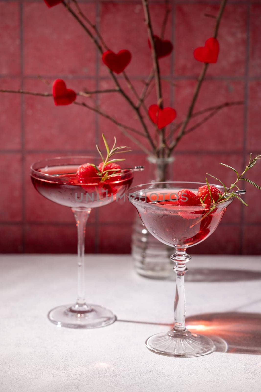Two elegant glasses filled with a vibrant pink cocktail, garnished with fresh raspberries and rosemary sprigs, create a romantic valentine's day atmosphere