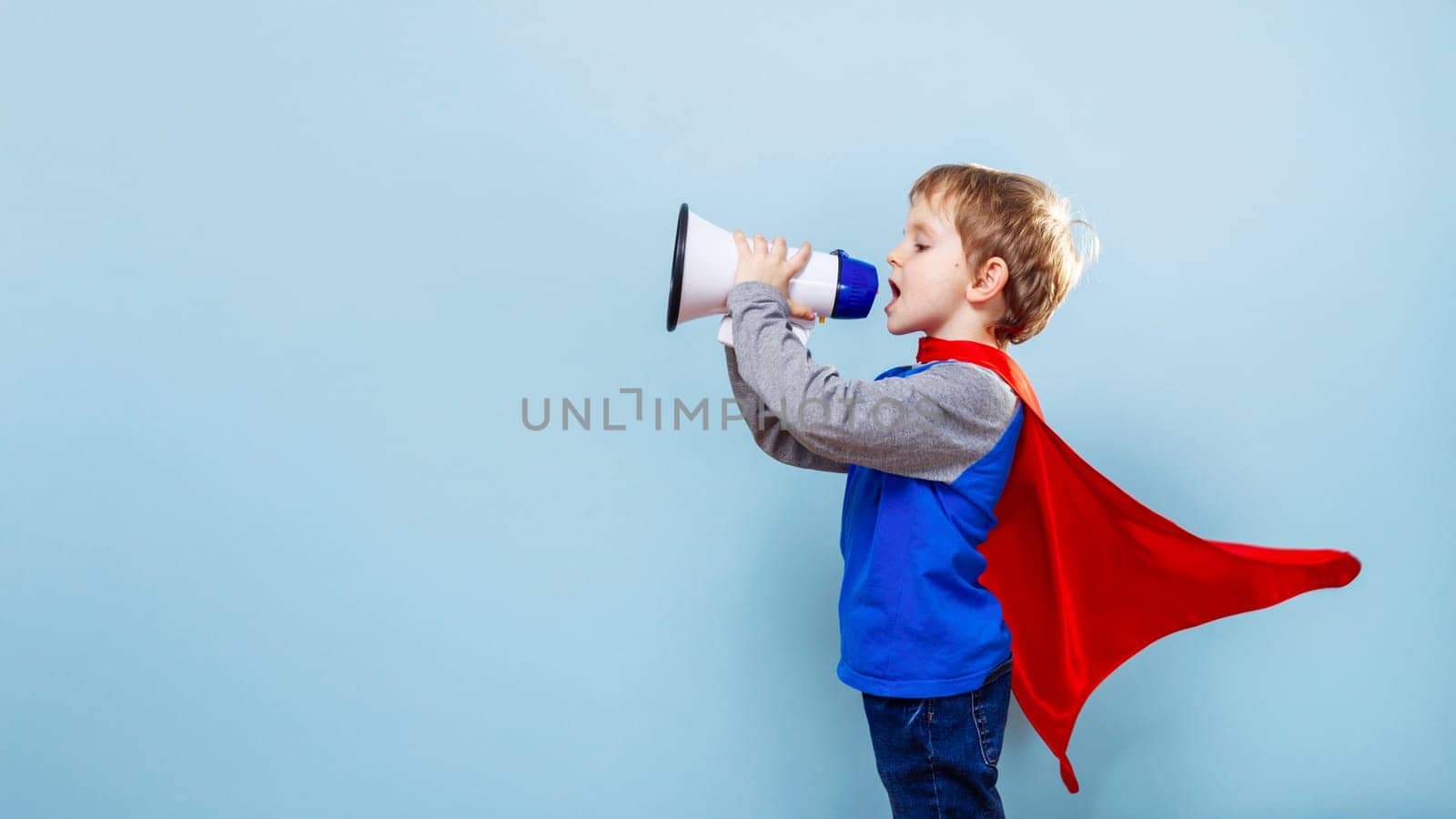 Young boy wearing a red cape and blue shirt holding a megaphone, shouting against a light blue background. The child is dressed as a superhero, pretending to give an announcement
