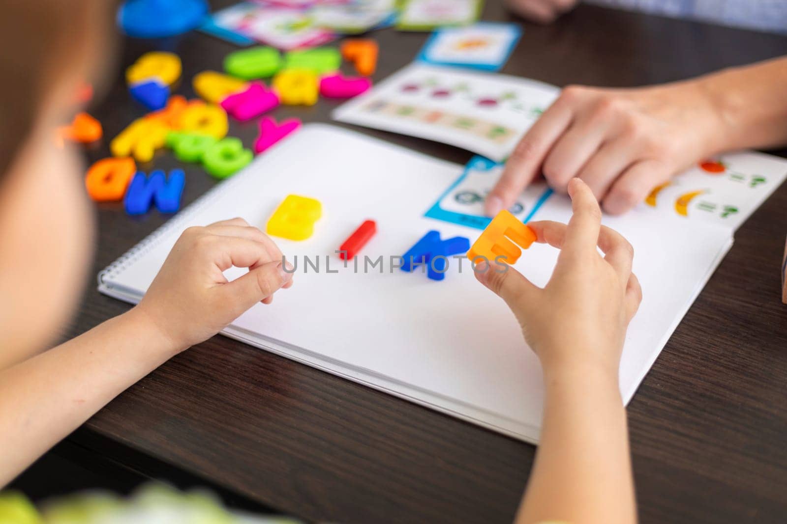 A child learns spelling using colorful plastic letters on a notebook, forming a word while an adult points at an educational activity sheet. Indoor home or classroom learning environment