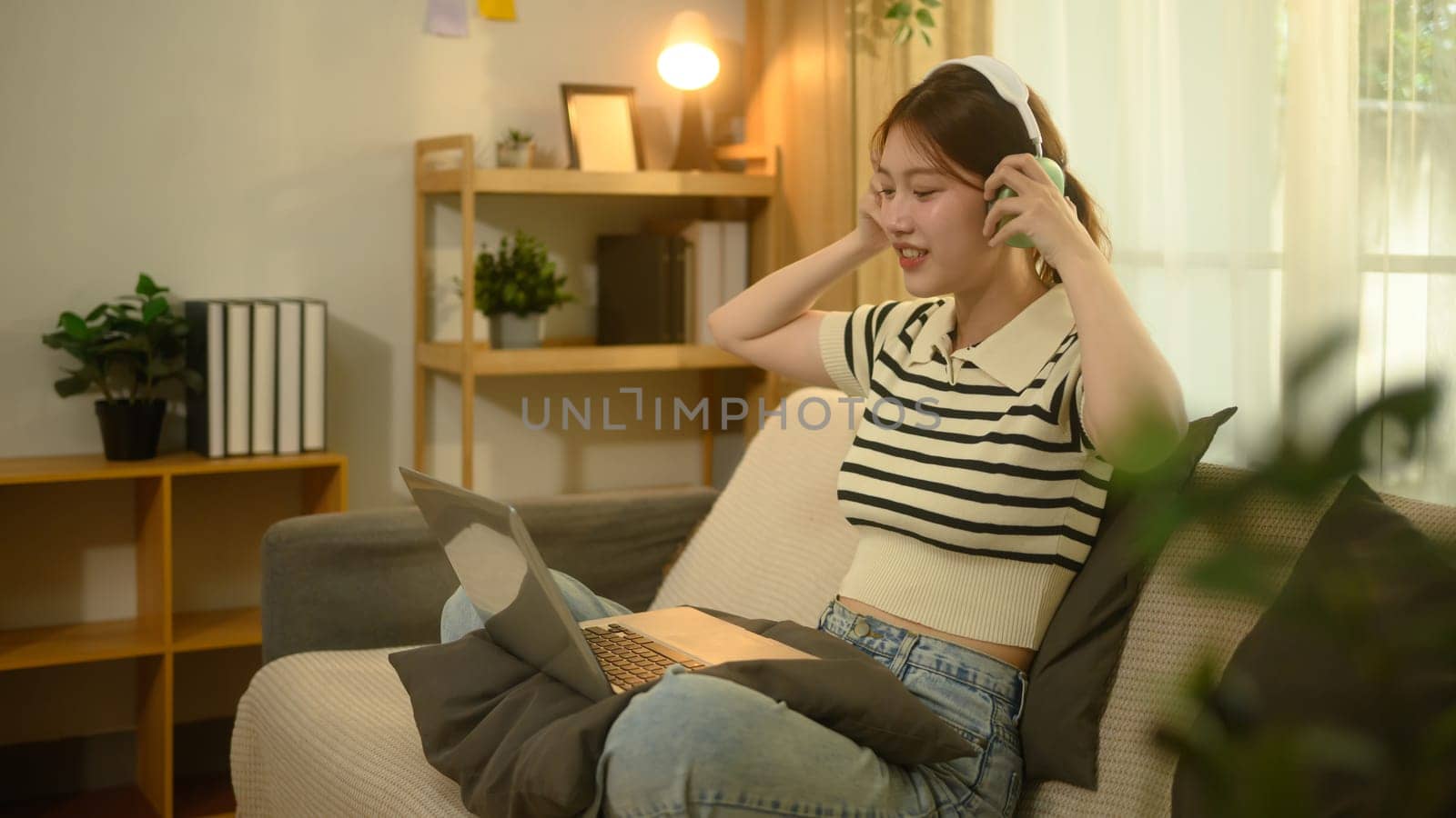 Young asian woman wearing wireless headphones and working on her laptop in living room.