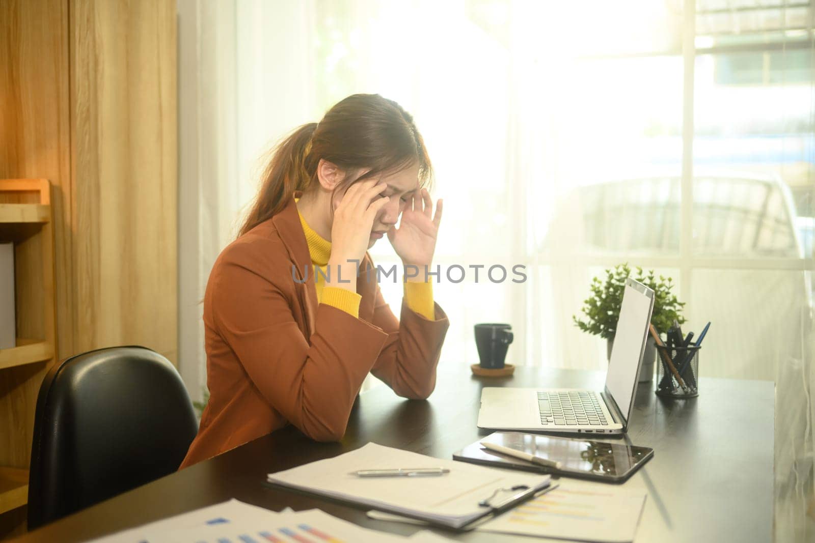 Young businesswoman sitting at her desk full of documents, holding her head in frustration.