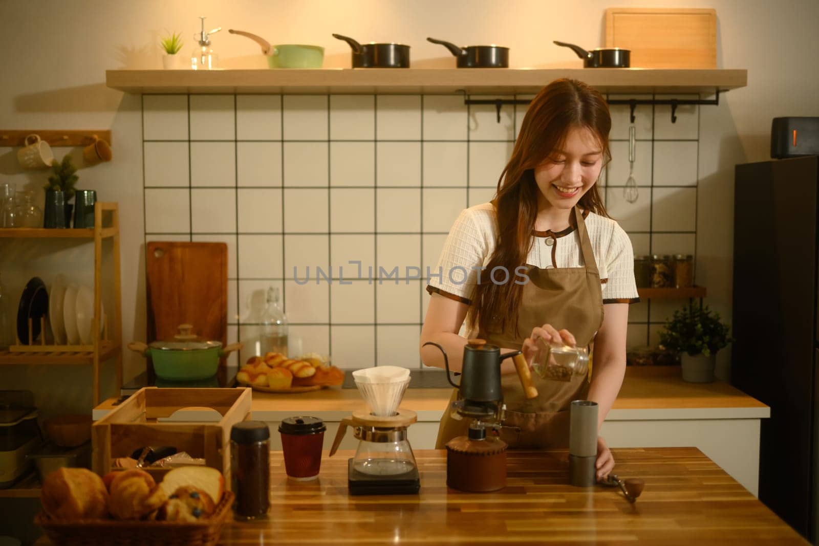 Woman in apron pouring coffee beans into a manual grinder on a wooden kitchen counter.