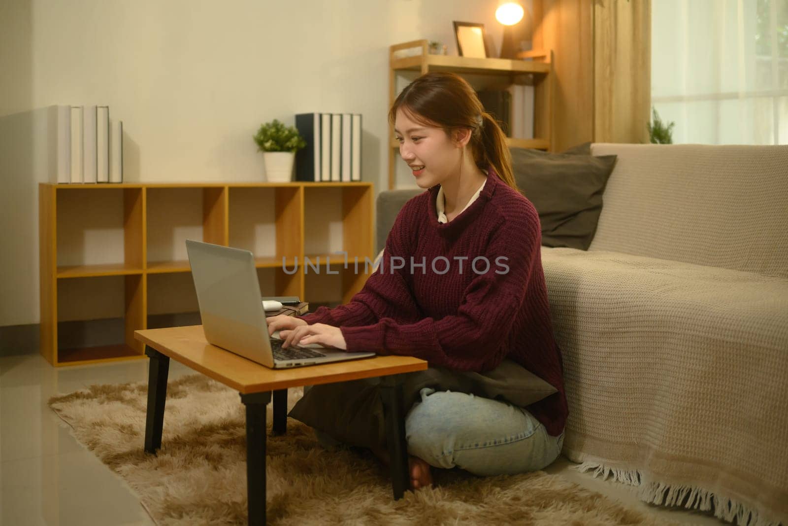 Smiling young woman in a maroon sweater using laptop in a cozy living room.