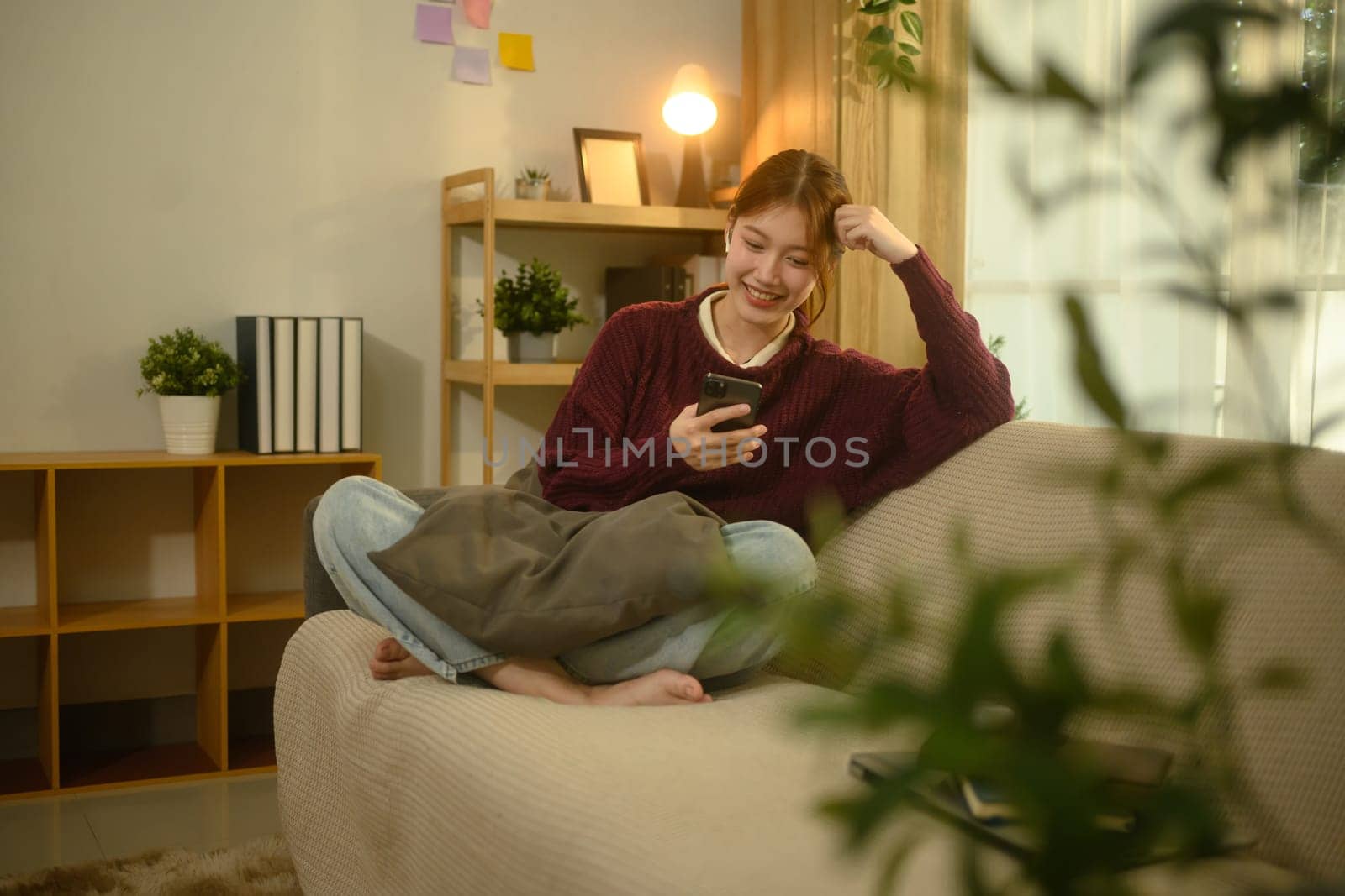 Smiling young asian woman enjoying social media on smartphone sitting on couch at home.
