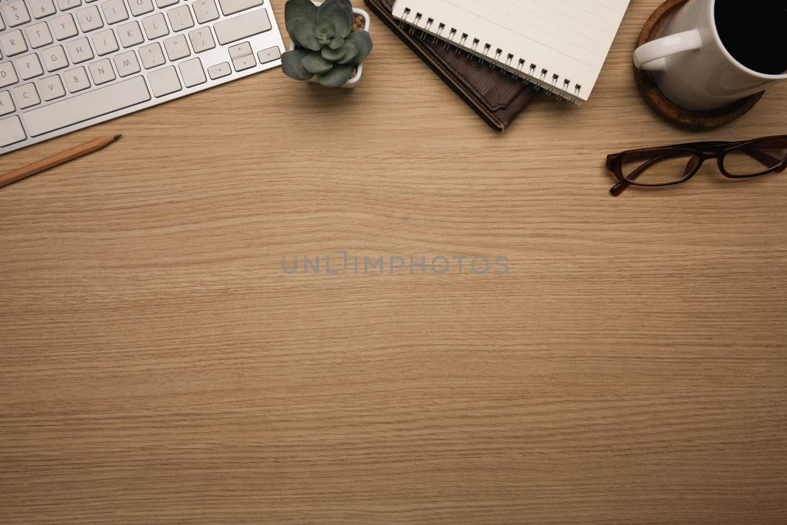 Top view shot of wooden desk with wireless keyboard, notebook, a cup of coffee and eyeglasses.