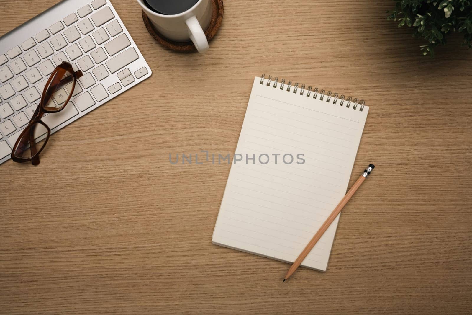 Blank spiral notebook, a cup of black coffee, eyeglasses, and wireless keyboard on wooden table.