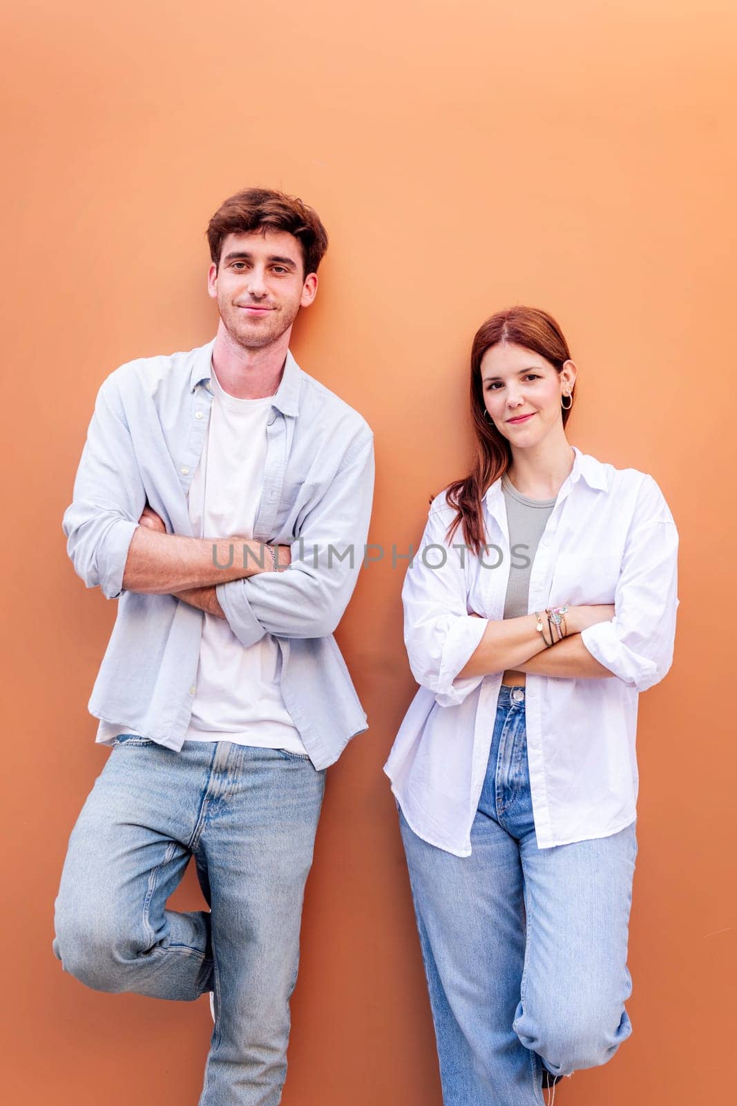 happy young couple of man and woman standing in front of orange wall looking at camera with arms crossed, concept of relationship and friendship