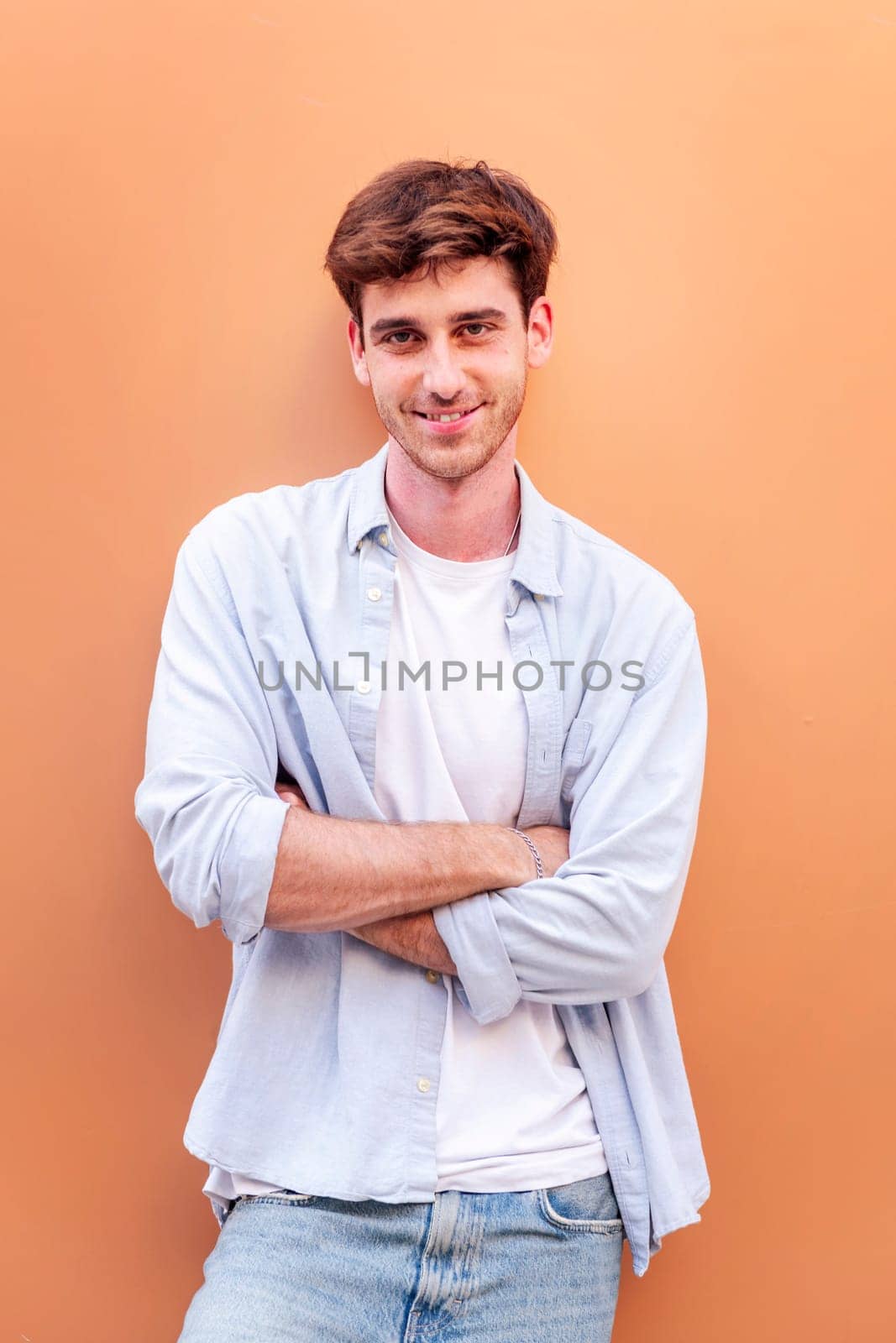 smiling young man standing in front of orange wall looking at camera with his arms crossed, he is wearing a white shirt and blue jeans