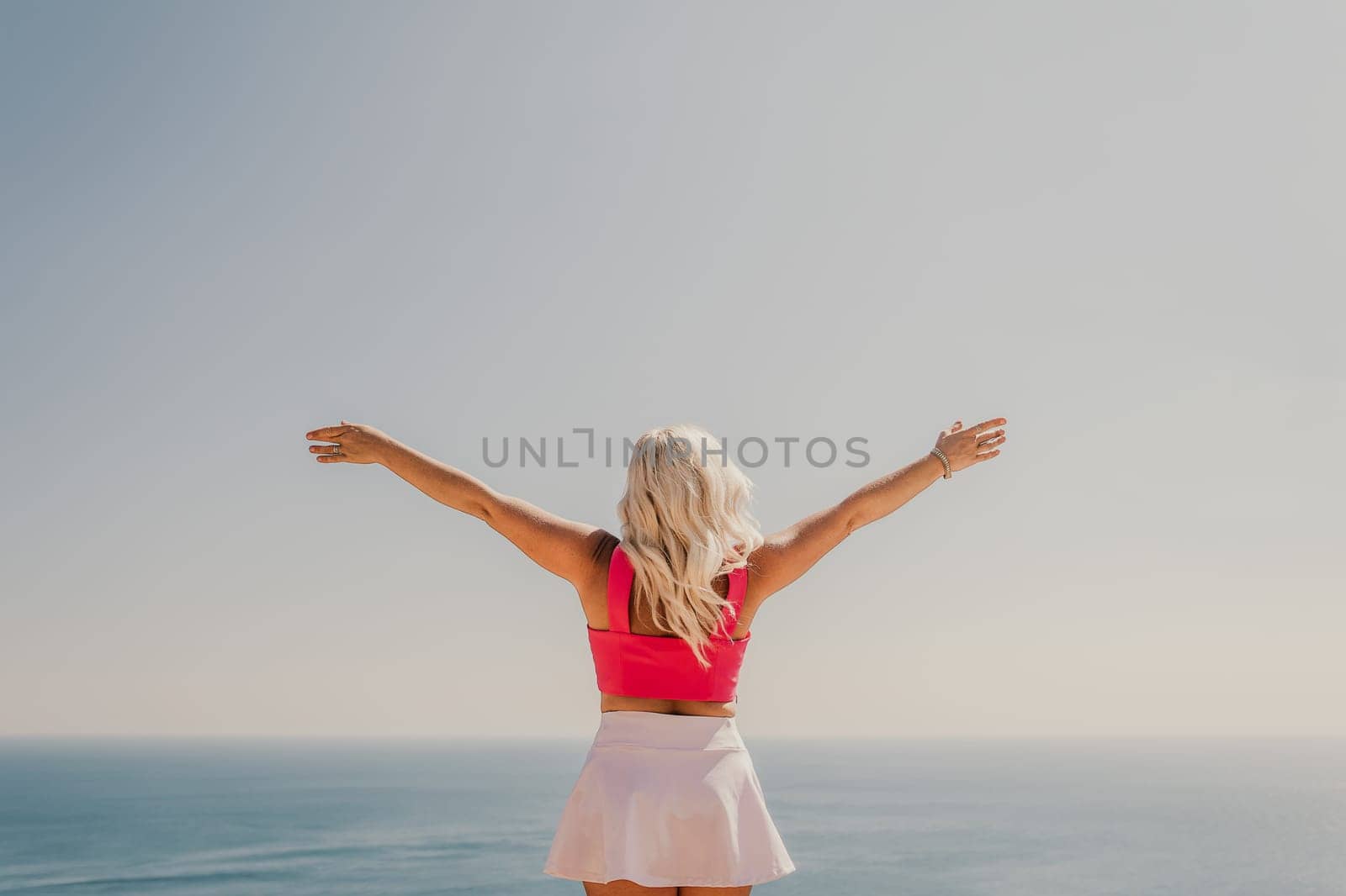 A woman in a pink top and white skirt is standing on a cliff overlooking the ocean. She is smiling and she is enjoying the view
