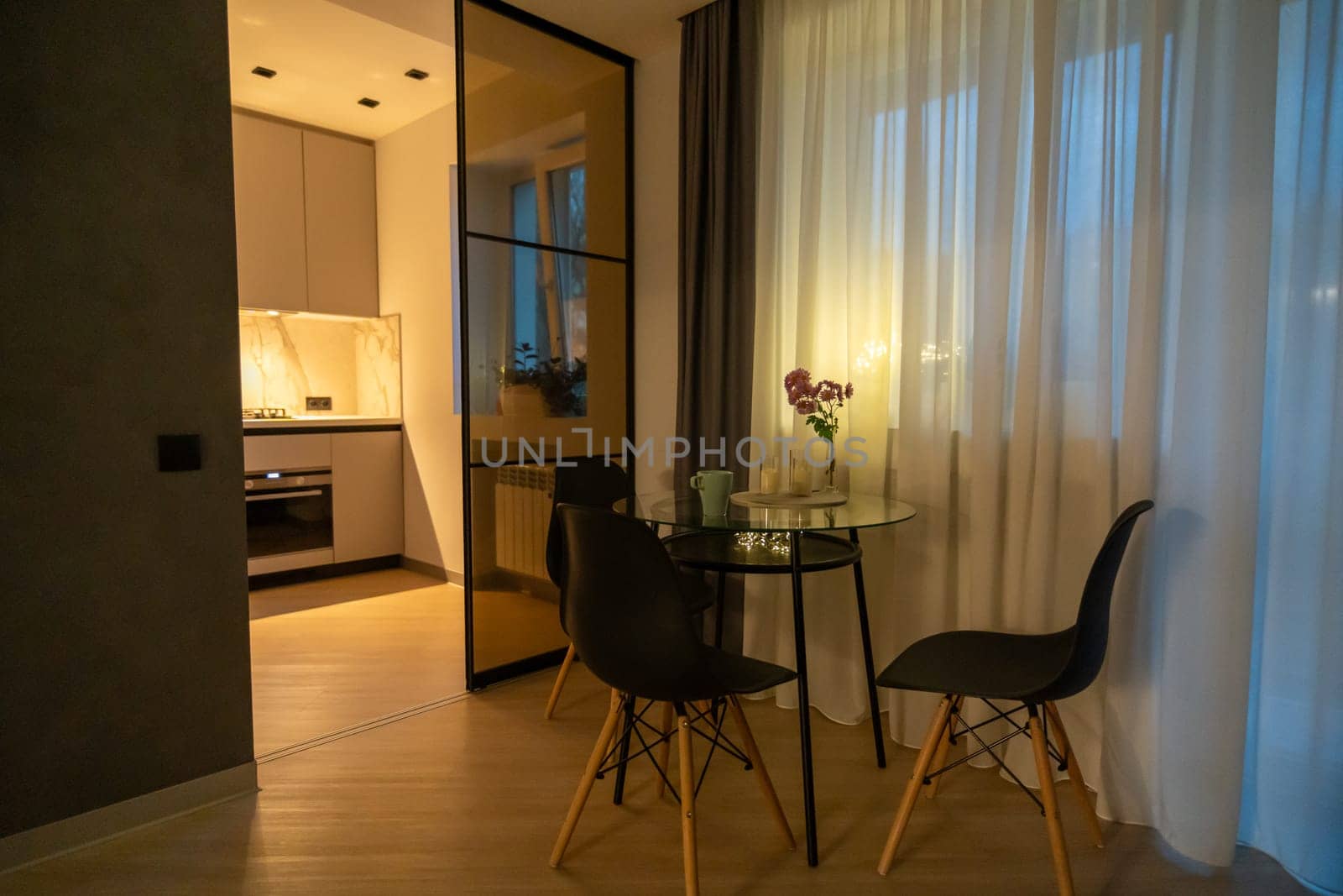 Modern apartment's dimly lit dining area. Glass-topped table with four black chairs sits near a glass-paned doorway leading to a kitchen. Sheer curtains frame a window.