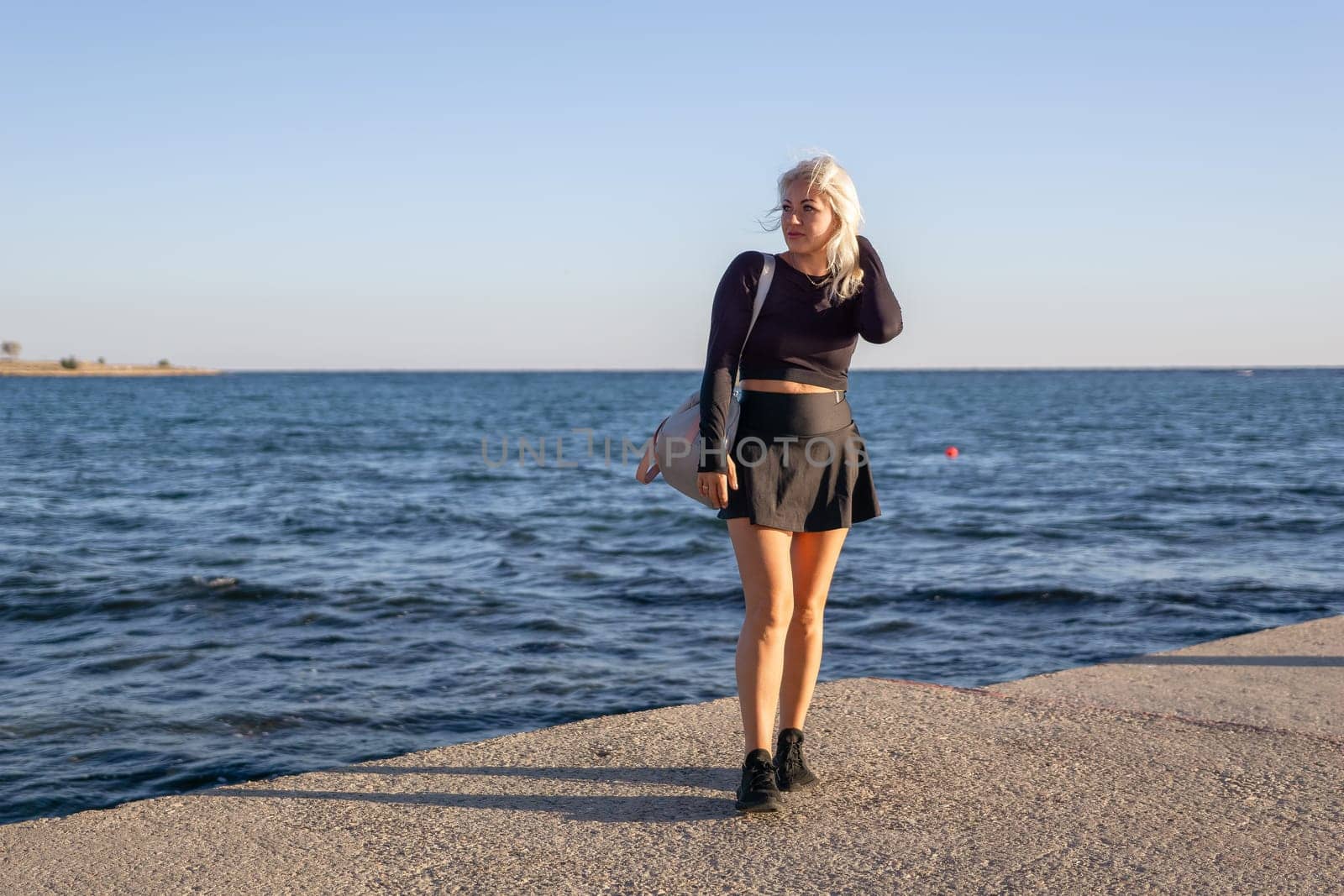 A blonde woman in a black skirt and top stands by the ocean, looking at the camera with a smile. She is holding a bag in her right hand.