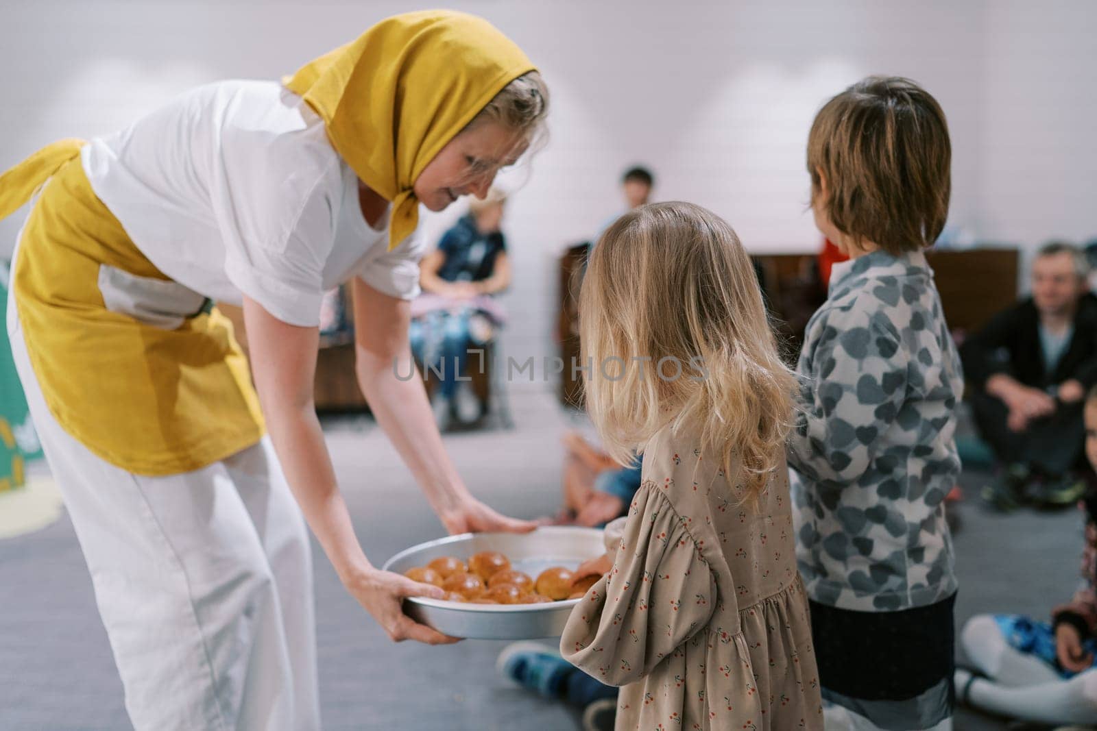Smiling actress distributes buns to children in kindergarten from a tray in her hands. High quality photo