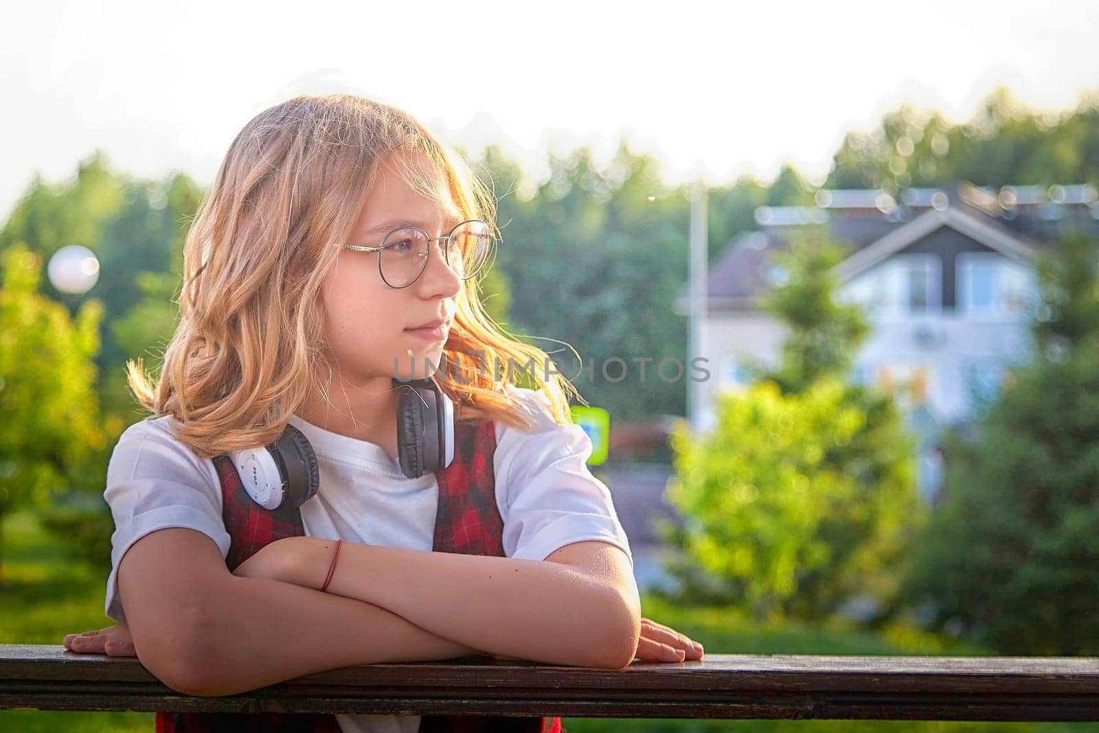 Girl with glasses and headphones leans on a railing, gazing thoughtfully at the lush greenery and homes nearby.