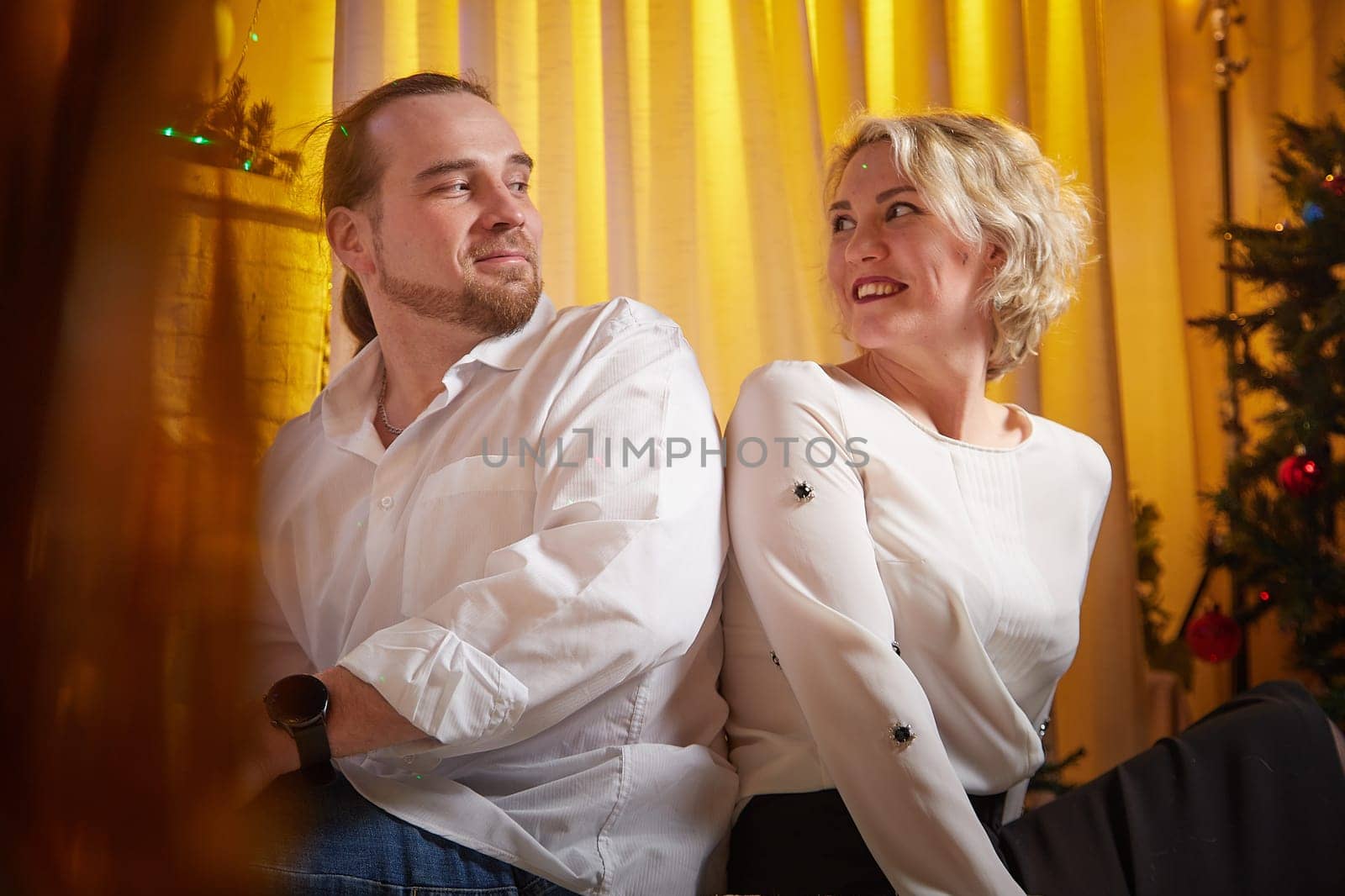 Couple shares a light-hearted moment while sitting beside a decorated Christmas tree in a cozy, festive atmosphere.