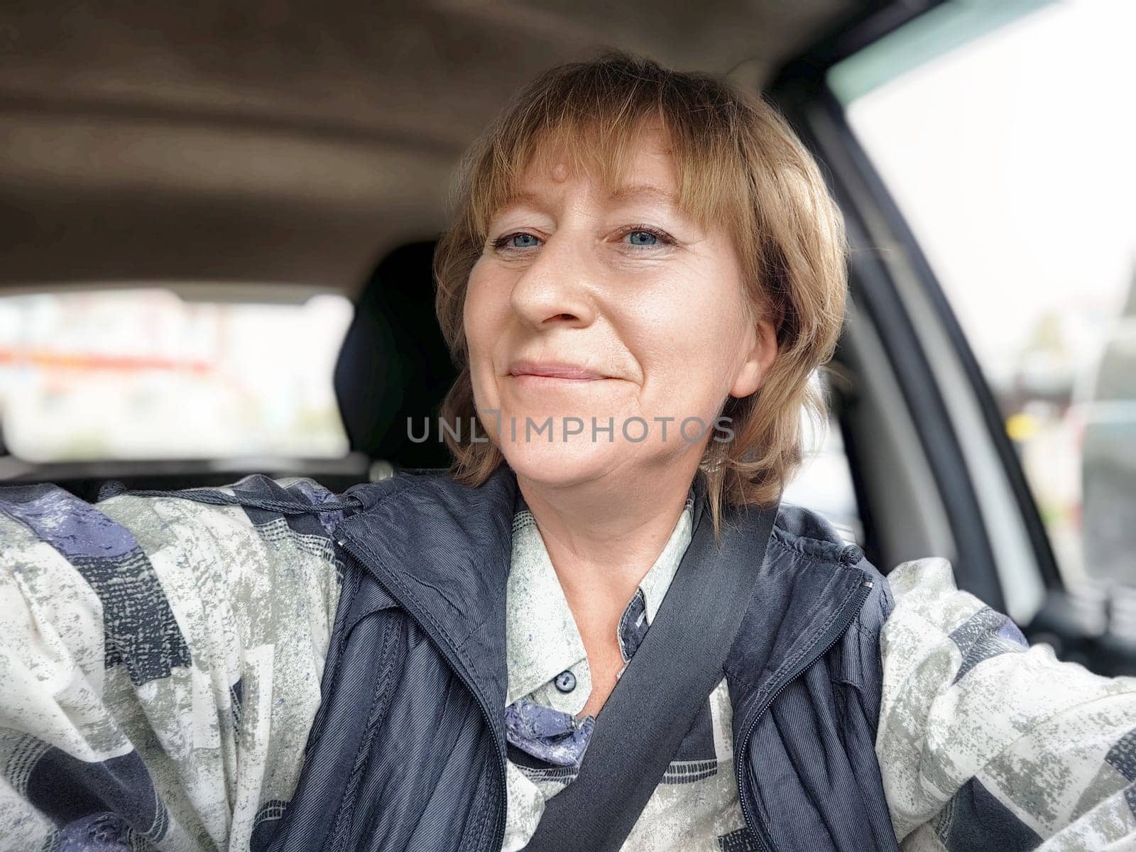 A middle-aged woman driver smiles while taking a selfie, capturing her joyful travel experience on the road.