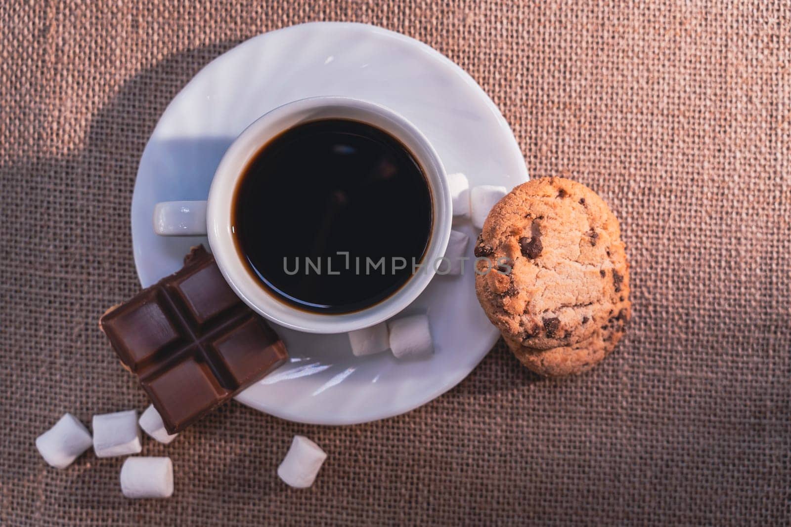 A top-down view of a cup of black coffee with chocolate, a cookie, and marshmallows on a white plate over a textured burlap background. Warm and cozy composition