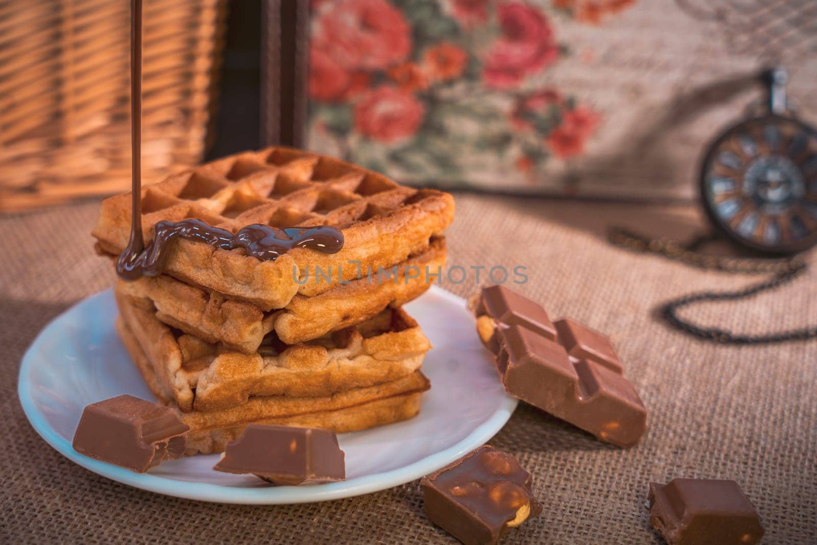 A stack of golden waffles with rich chocolate sauce dripping on top, surrounded by chocolate pieces, set on a white plate with a vintage pocket watch in the background. Warm and indulgent scene