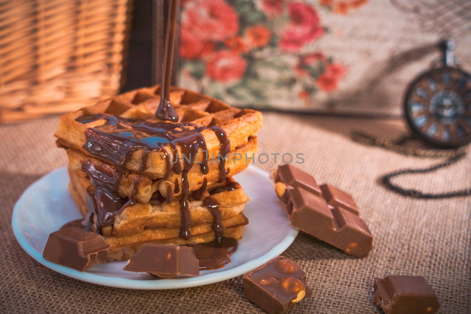 A stack of golden waffles with rich chocolate sauce dripping on top, surrounded by chocolate pieces, set on a white plate with a vintage pocket watch in the background. Warm and indulgent scene