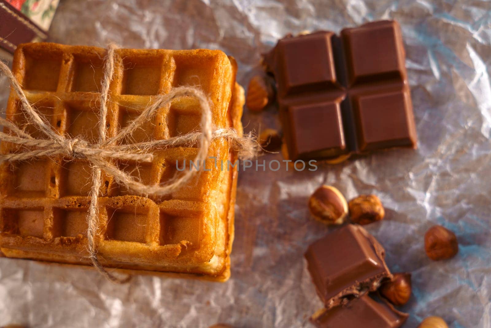 A stack of golden waffles tied with twine, accompanied by broken chocolate pieces and hazelnuts on crinkled parchment paper. A warm and rustic food composition