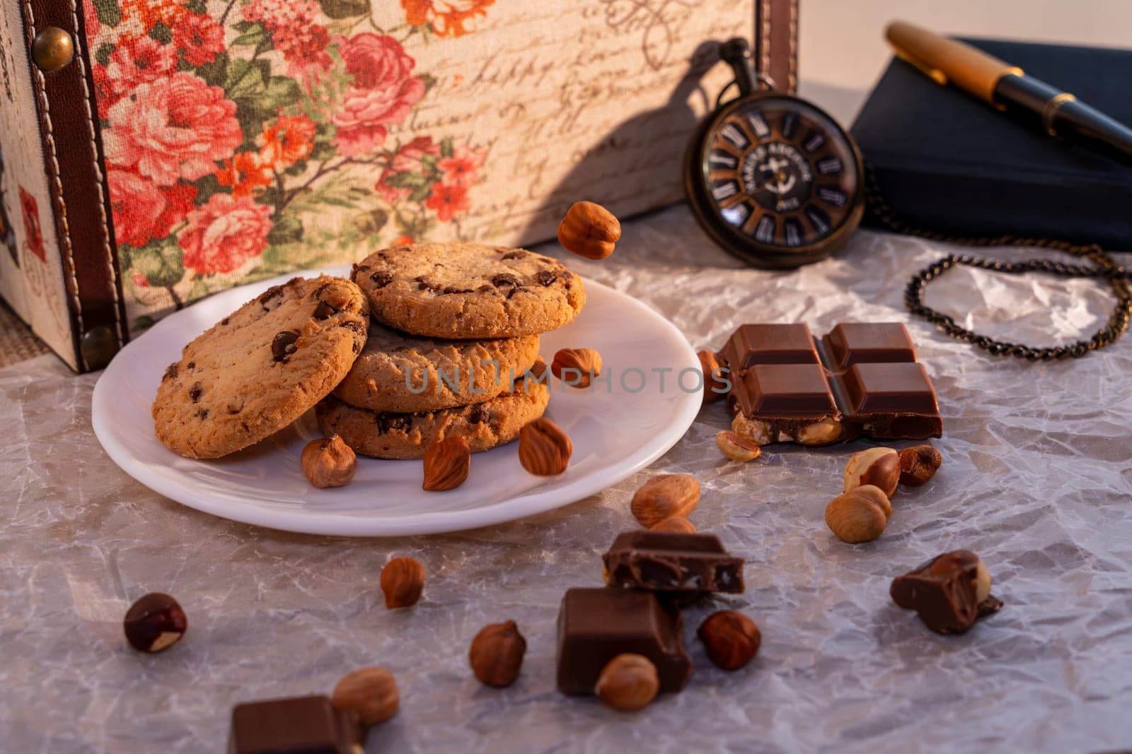 A plate of chocolate chip cookies with broken chocolate pieces and hazelnuts on crinkled parchment paper, accompanied by a vintage pocket watch, notebook, and floral box. Cozy and nostalgic setting