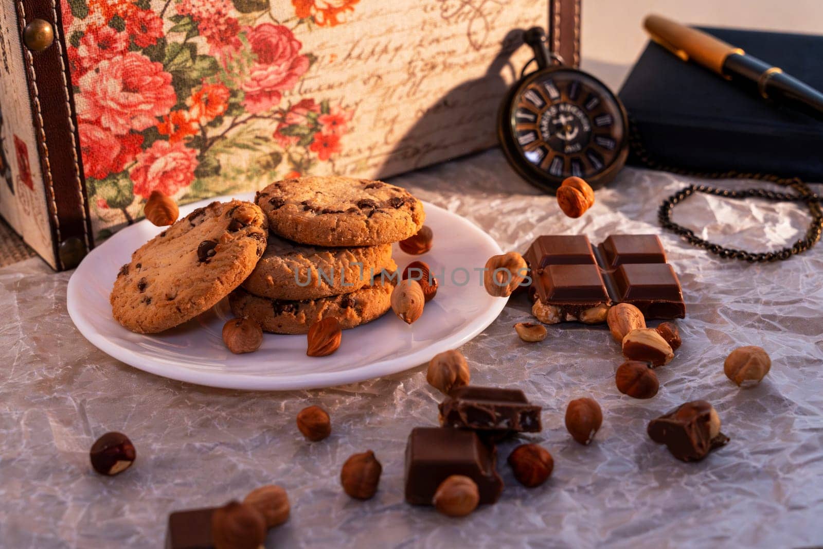 A plate of chocolate chip cookies with broken chocolate pieces and hazelnuts on crinkled parchment paper, accompanied by a vintage pocket watch, notebook, and floral box. Cozy and nostalgic setting