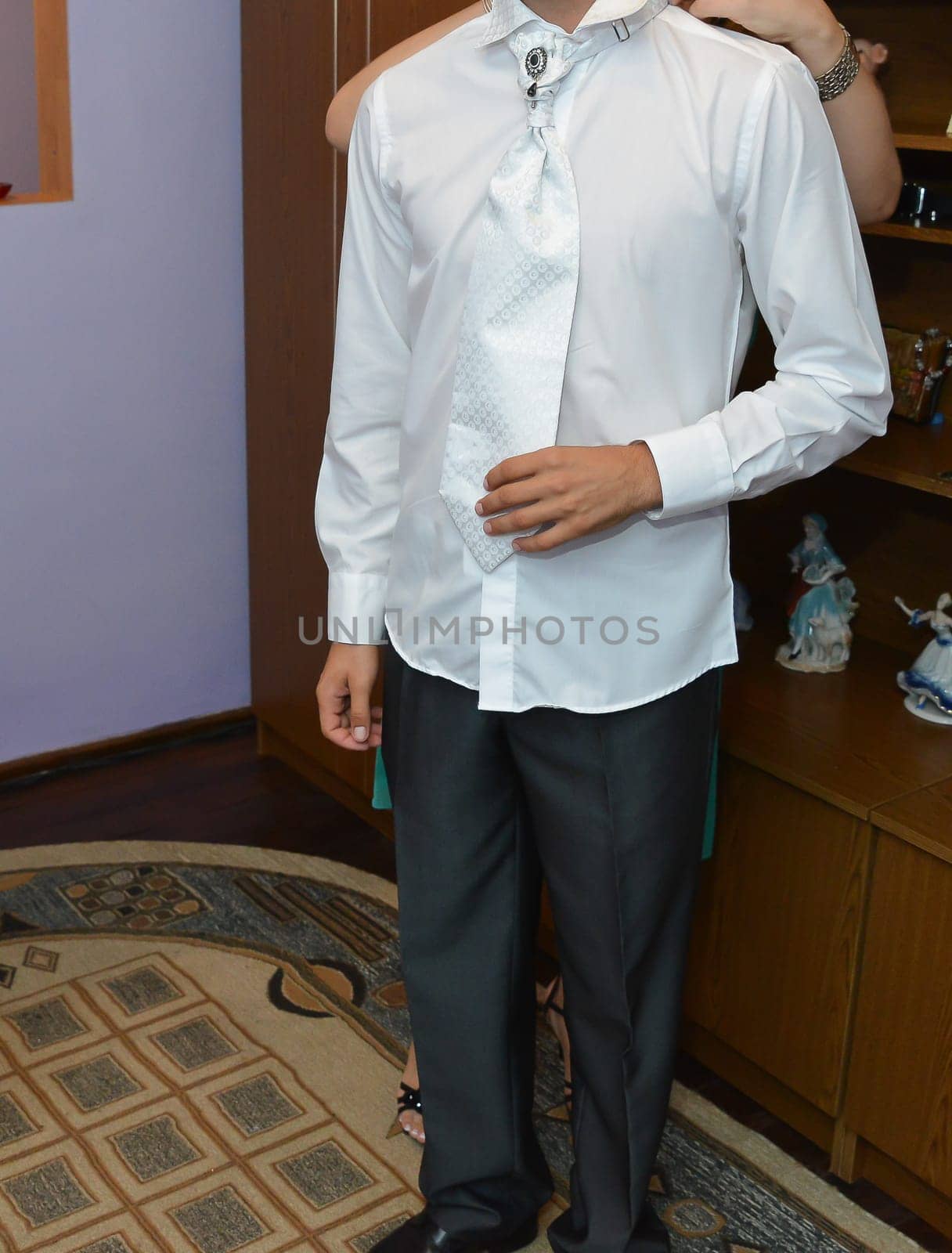A man dressed in formal attire, wearing a crisp white shirt, a patterned tie, and dark trousers, getting ready for a memorable event with a helping hand in the background.