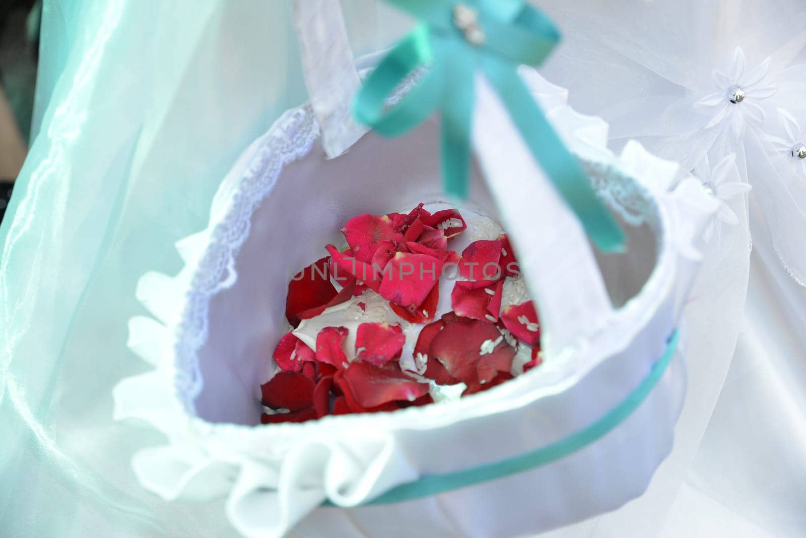 A beautifully decorated white basket adorned with lace and a teal ribbon, filled with red rose petals, symbolizing love and celebration at a special event.