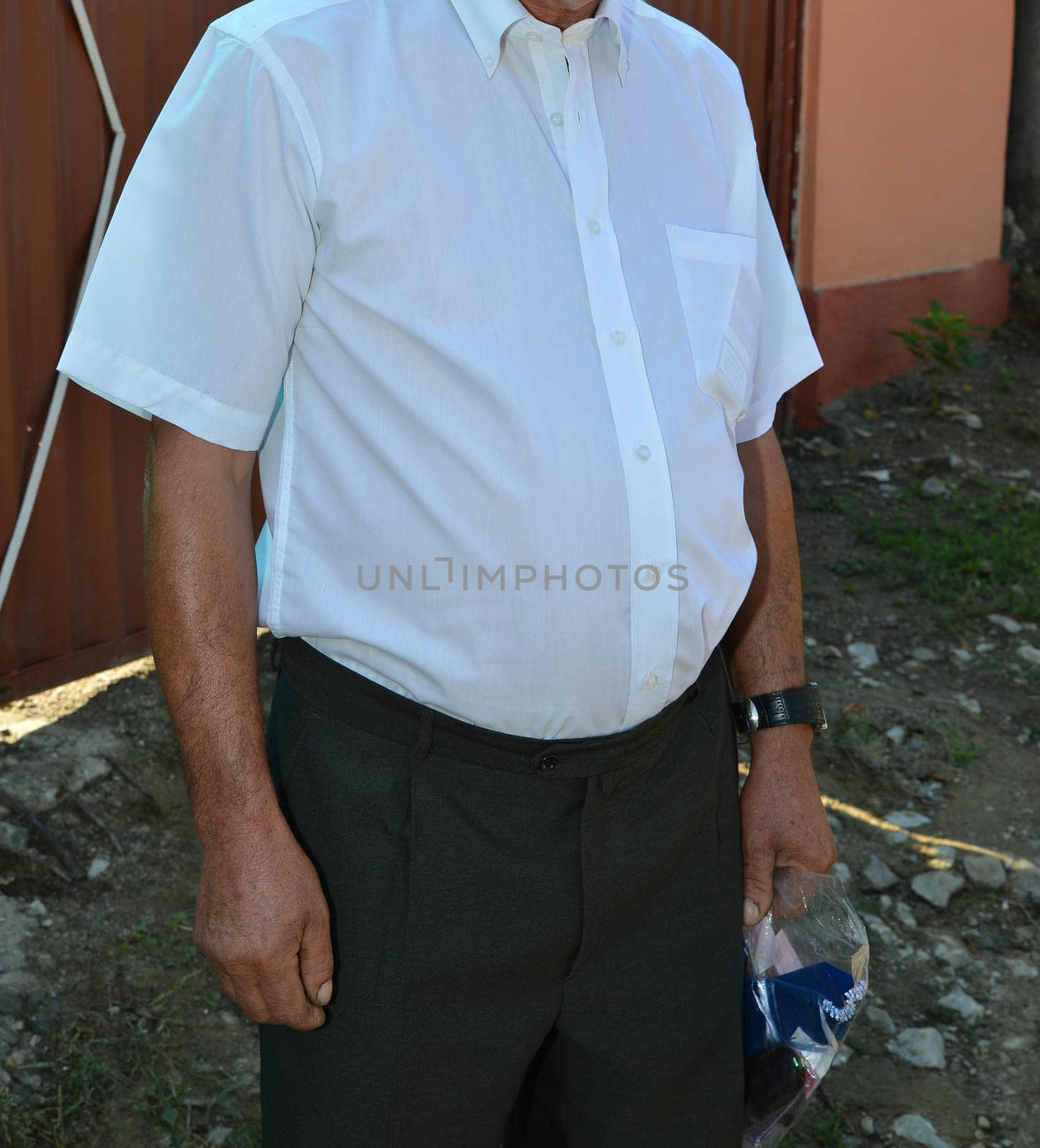 An elderly man dressed in a neatly pressed white short-sleeved shirt and dark trousers, standing outdoors with a plastic bag in hand, exuding a formal yet relaxed presence.