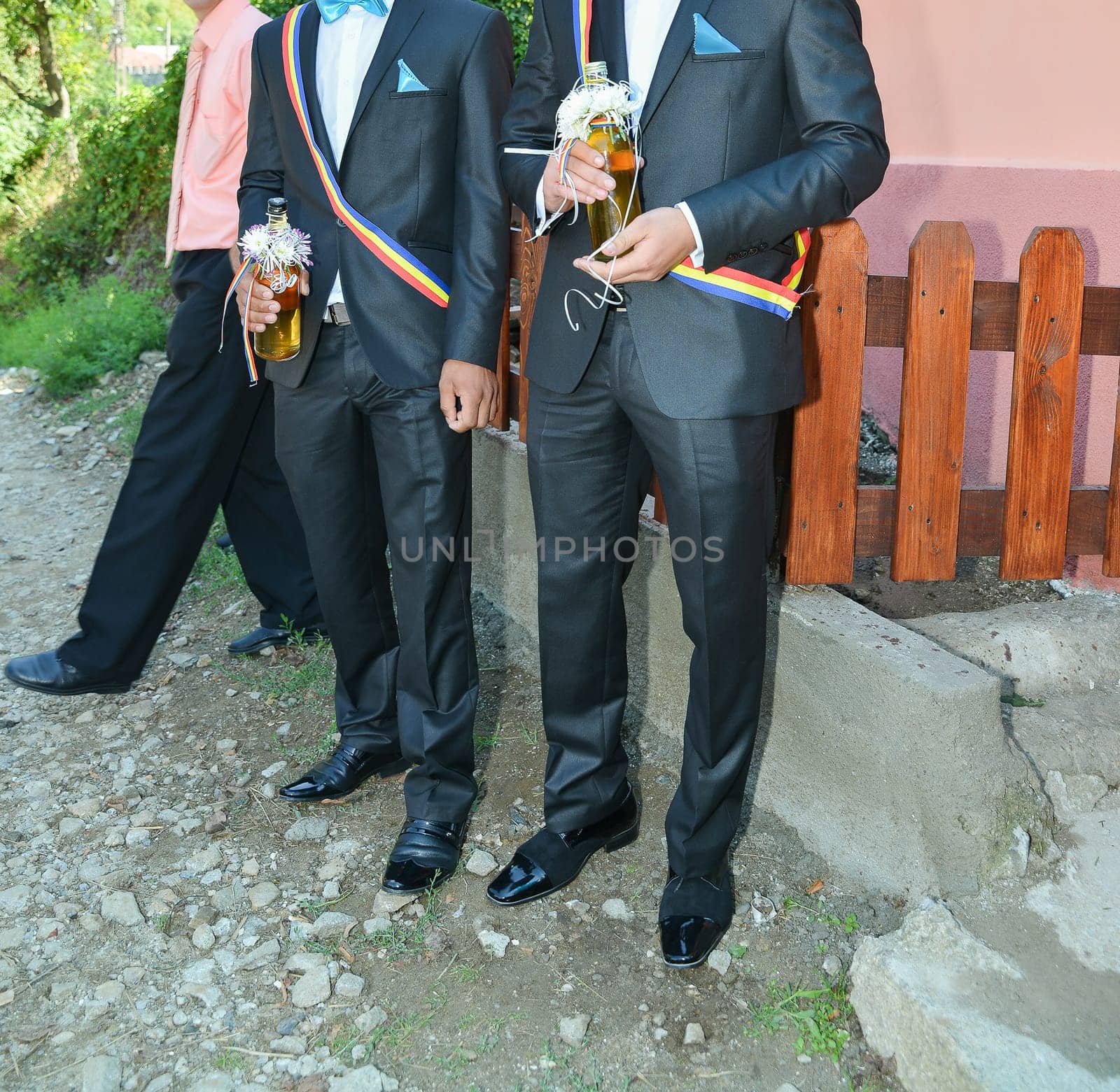 Two men in formal suits hold drinks, displaying vibrant sashes and stylish shoes, embodying the spirit of celebration.