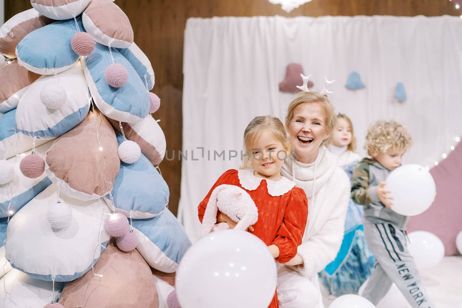 Little girl stands next to a smiling actress with small deer antlers near a toy Christmas tree on stage. High quality photo