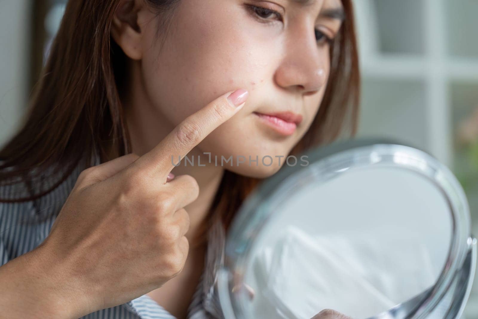 A close-up of a woman examining her skin in the mirror, pointing at a blemish, reflecting the challenges of her beauty regime.