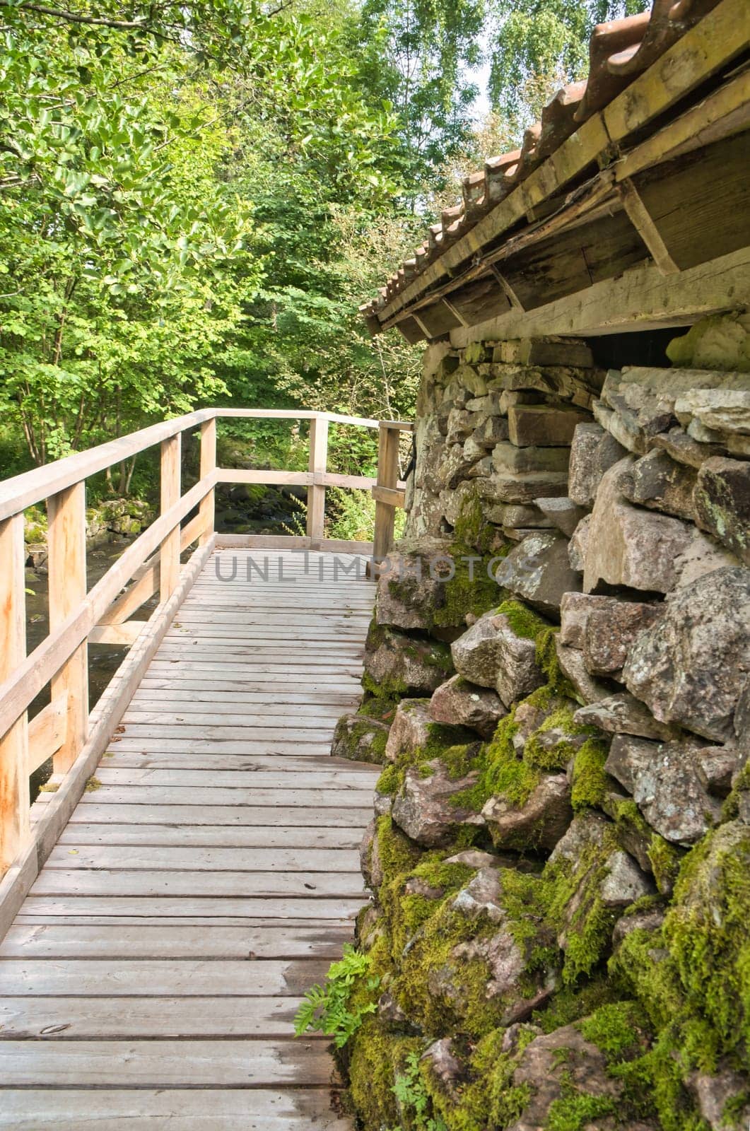 Old watermill surrounded by green nature. A small stream flows over moss-covered stones, surrounded by trees. Idyllic landscape with rustic wooden architecture.
