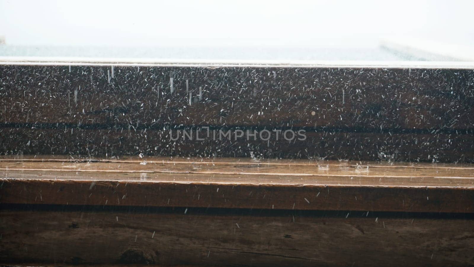 Close-up of a wooden window frame with rain and snow falling heavily against the glass during a winter storm, creating a cozy and dramatic atmosphere