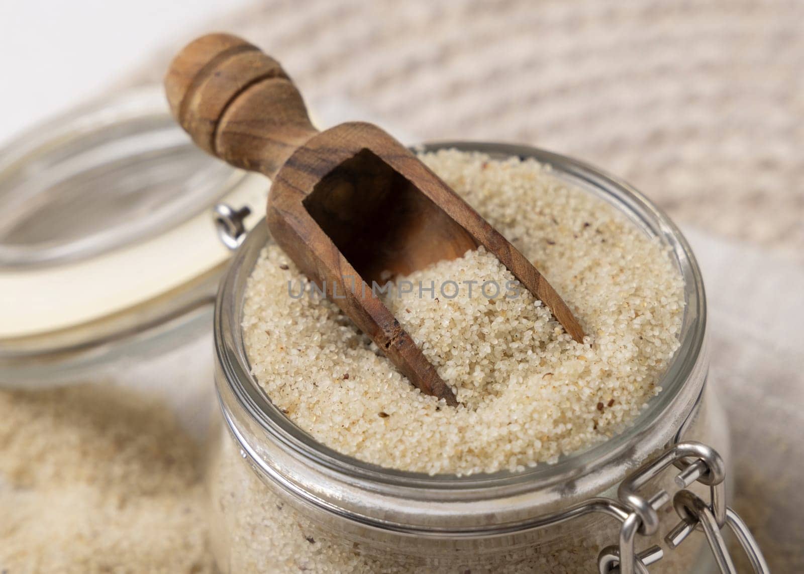 Glass jar of raw uncooked fonio seeds with a wooden scoop on a napkin on white table closeup. African ancient pseudocereal. Organic super food. Healthy food concept. Gluten free alternative seeds