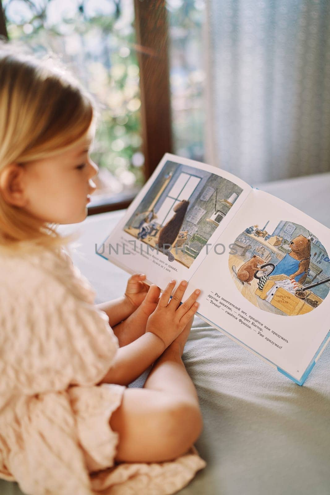 Little girl reads a picture book about a beaver workshop while sitting on her bed. Side view. High quality photo