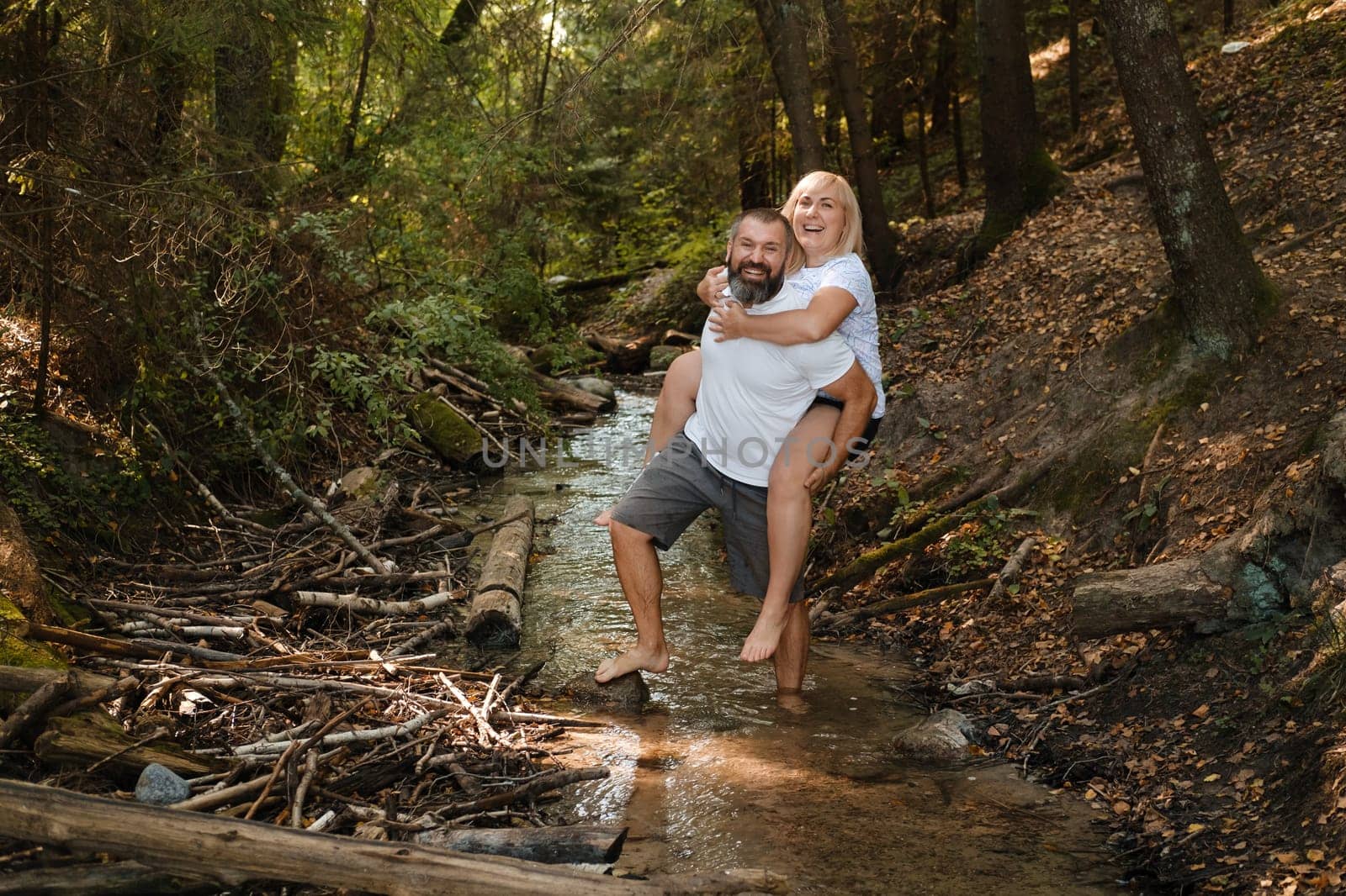 A joyful couple engages in a playful embrace, with the man giving a piggyback ride to the woman in a shallow stream surrounded by lush greenery, creating a cheerful and relaxed atmosphere.
