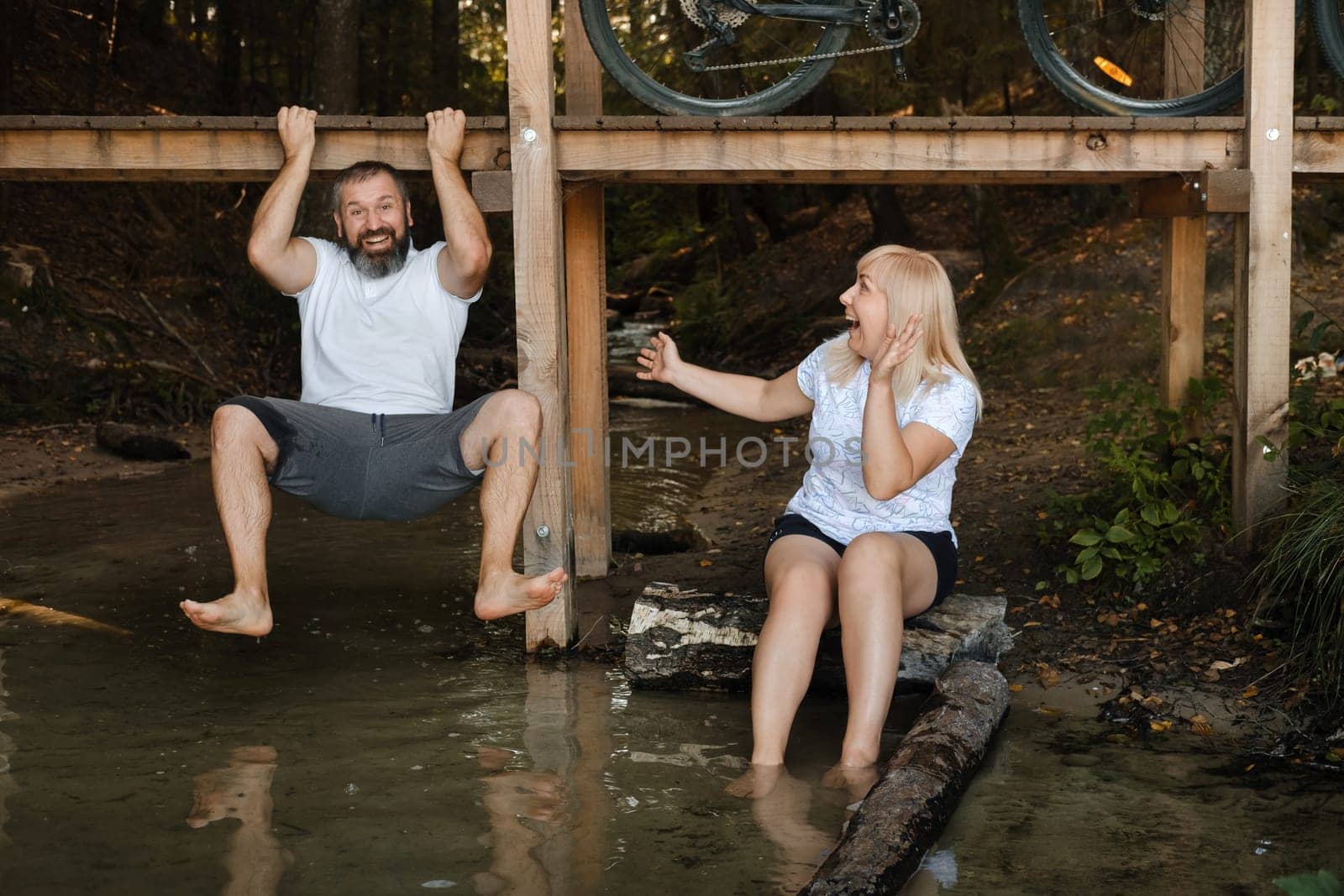 Two friends are having fun by the water on a sunny day. One is playfully hanging from a wooden platform while the other sits on a log, both smiling and enjoying the moment in nature.