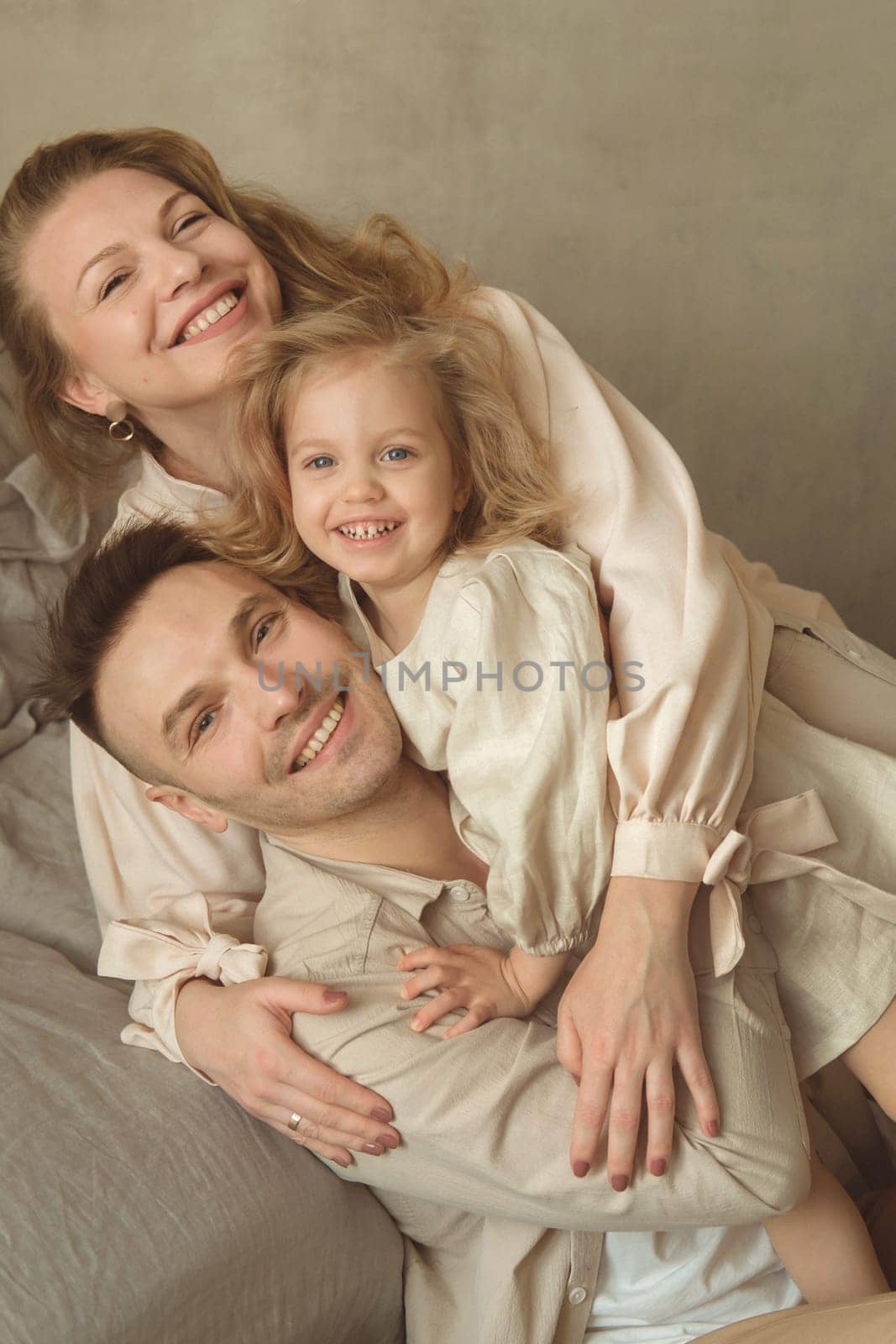 A joyful family of three enjoys a tender moment in a soft-toned indoor space. They are embracing each other with smiles, radiating affection and happiness together.
