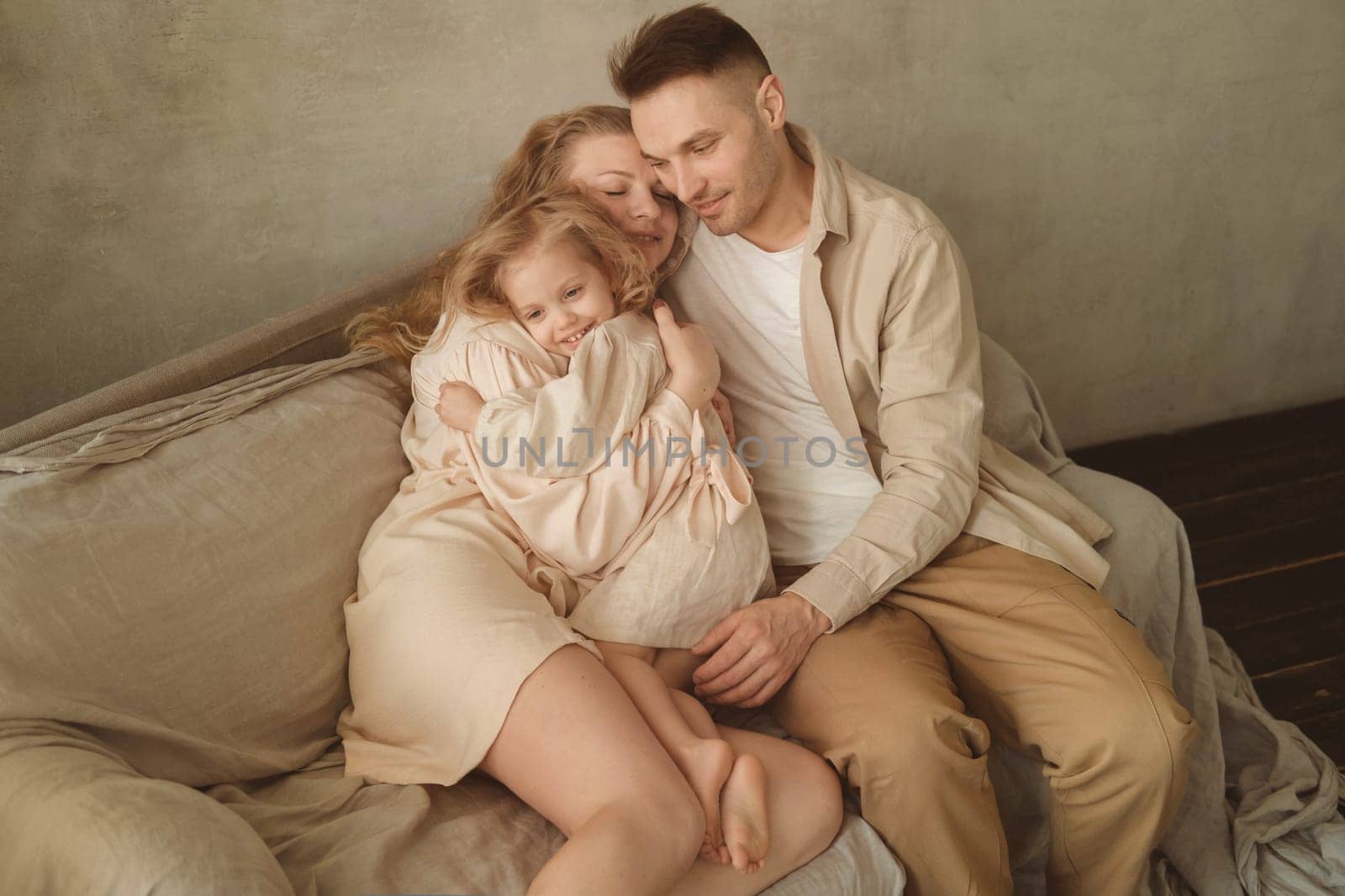 A mother, father, and young daughter embrace affectionately on a comfortable couch, enjoying a tender moment in their softly lit living room while dressed in relaxed clothing.