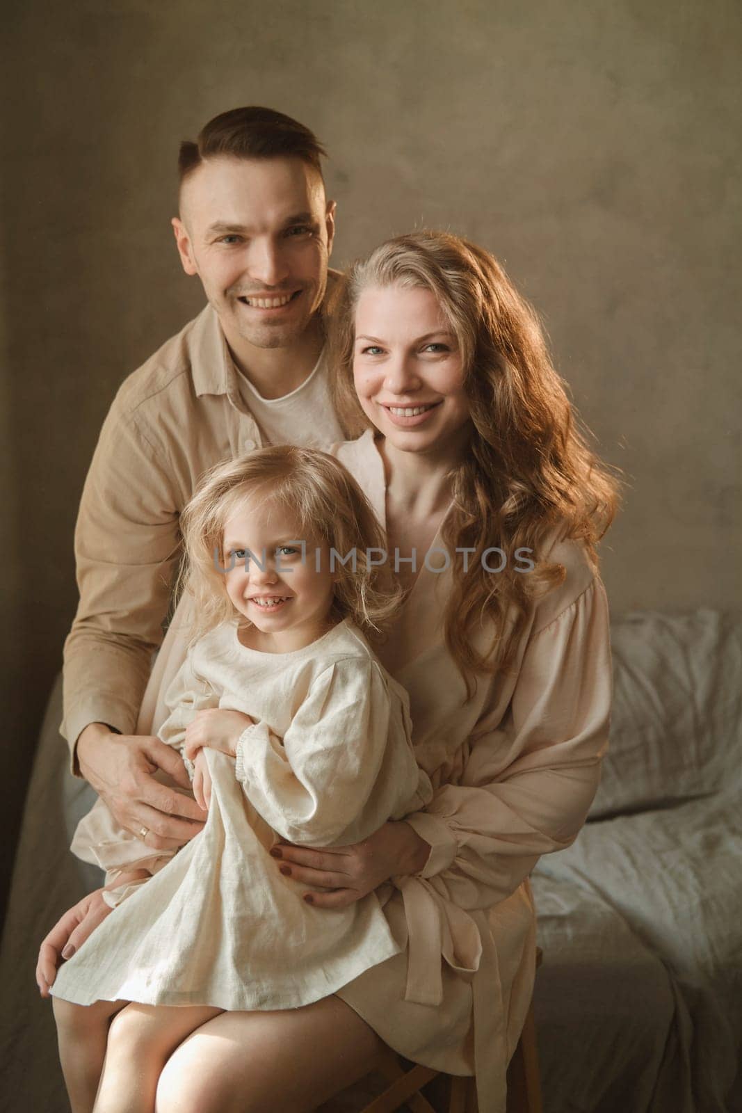 A happy family poses together in a warm and inviting indoor space, filled with soft light. The parents embrace their smiling child, showcasing love and togetherness in the moment.