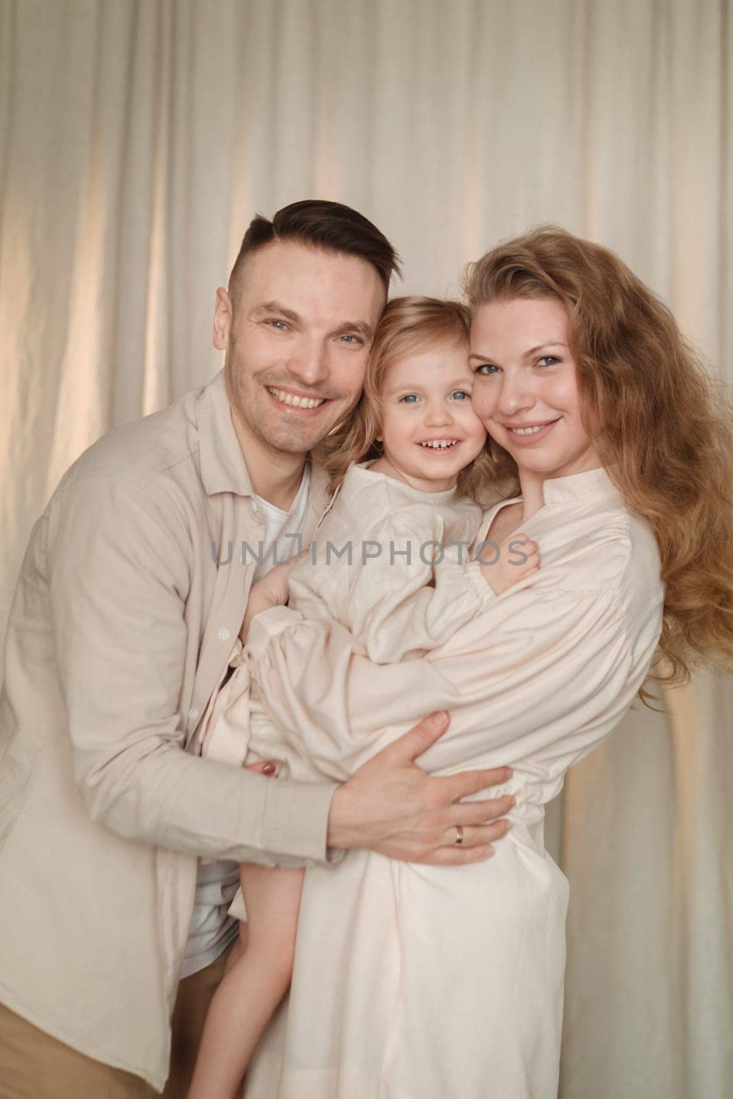 A happy family of three stands closely together, smiling warmly. The parents embrace their child, who is cheerful and playful. Soft lighting creates a warm atmosphere in the background.