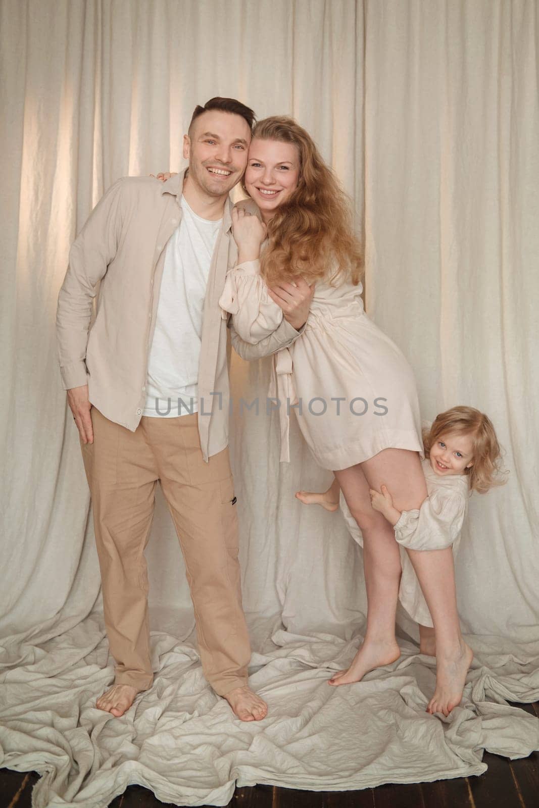 A cheerful family stands together in a soft-lit room. Parents smile warmly while their child playfully peeks from behind. Their casual outfits create a relaxed atmosphere filled with affection.